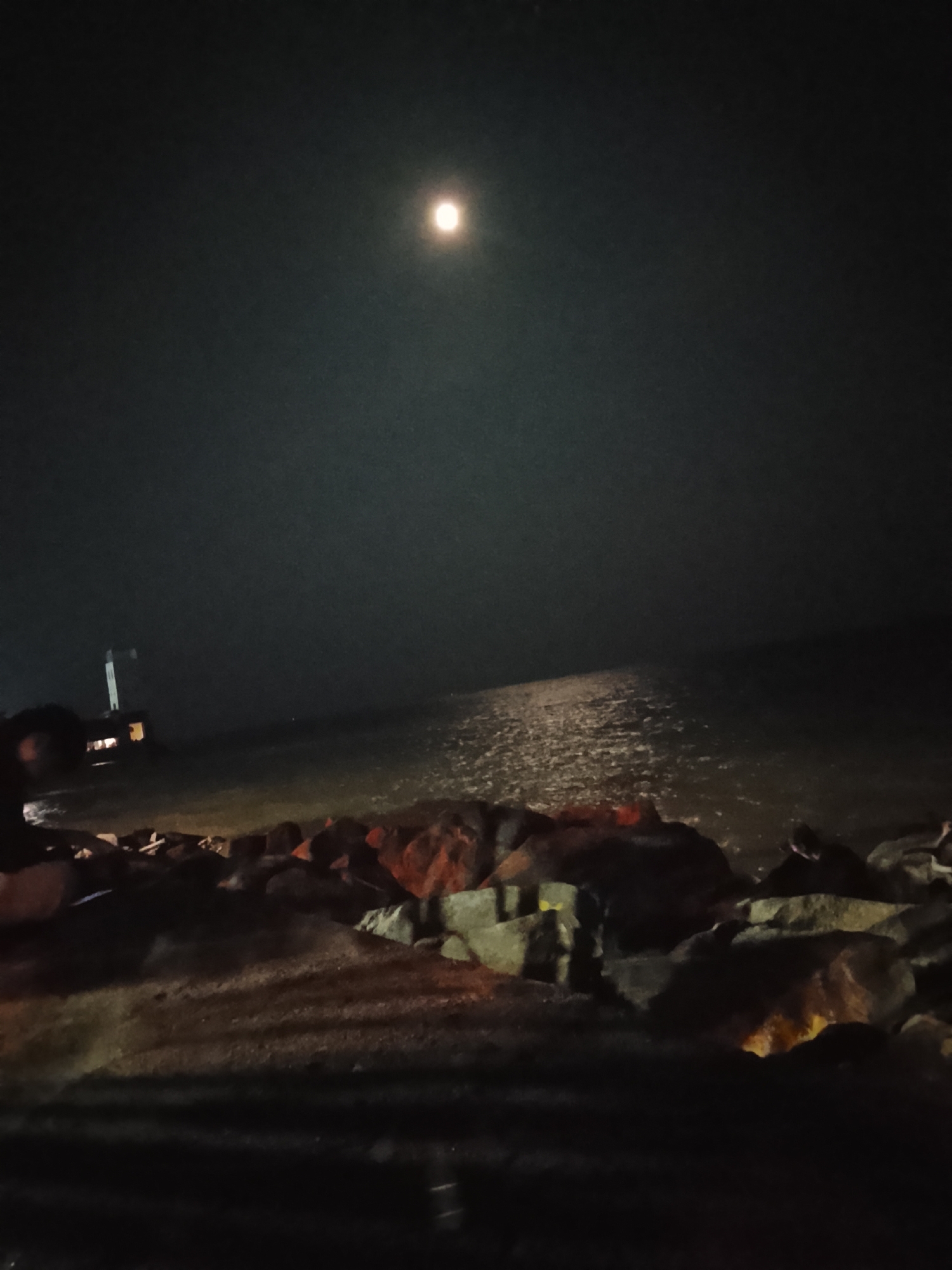 The image depicts a nighttime scene at the waterfront in Tanjung Balai Karimun, Indonesia. The sky is dark with a prominent bright moon positioned high and slightly to the left of the frame. A reflection of the moon shimmers on the dark water. In the foreground, a rocky shore is visible, with some of the rocks appearing to be illuminated by artificial lights, casting a reddish glow. To the left, a structure resembling a pier or a small building with lights is partially submerged in the water. The overall atmosphere is serene and tranquil, with the natural light of the moon dominating the scene. There are no people clearly visible in the image.
