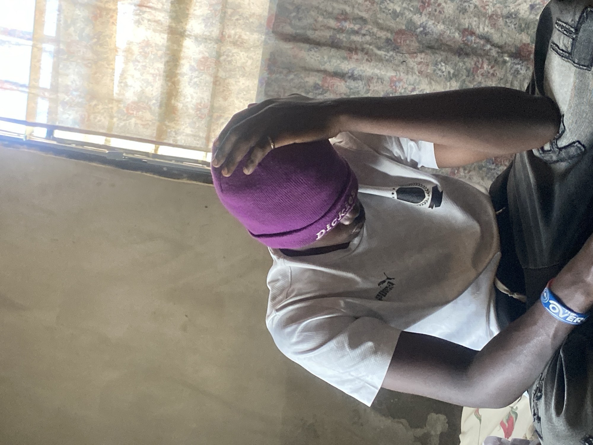 The media file shows a person wearing a white t-shirt and a purple beanie. The person's hand is resting on their head. The background is a window with curtains. There is also a patterned fabric, possibly a bedspread or blanket, visible. A blue wristband with white text that appears to say "OVER" is worn on the person's wrist. A small thermometer is visible in the person's mouth. The lighting suggests it may be daytime, with light coming from the window. The scene appears to be indoors, possibly a bedroom. The person's posture and hand on their head might suggest discomfort or contemplation. The overall impression is an intimate, candid moment.