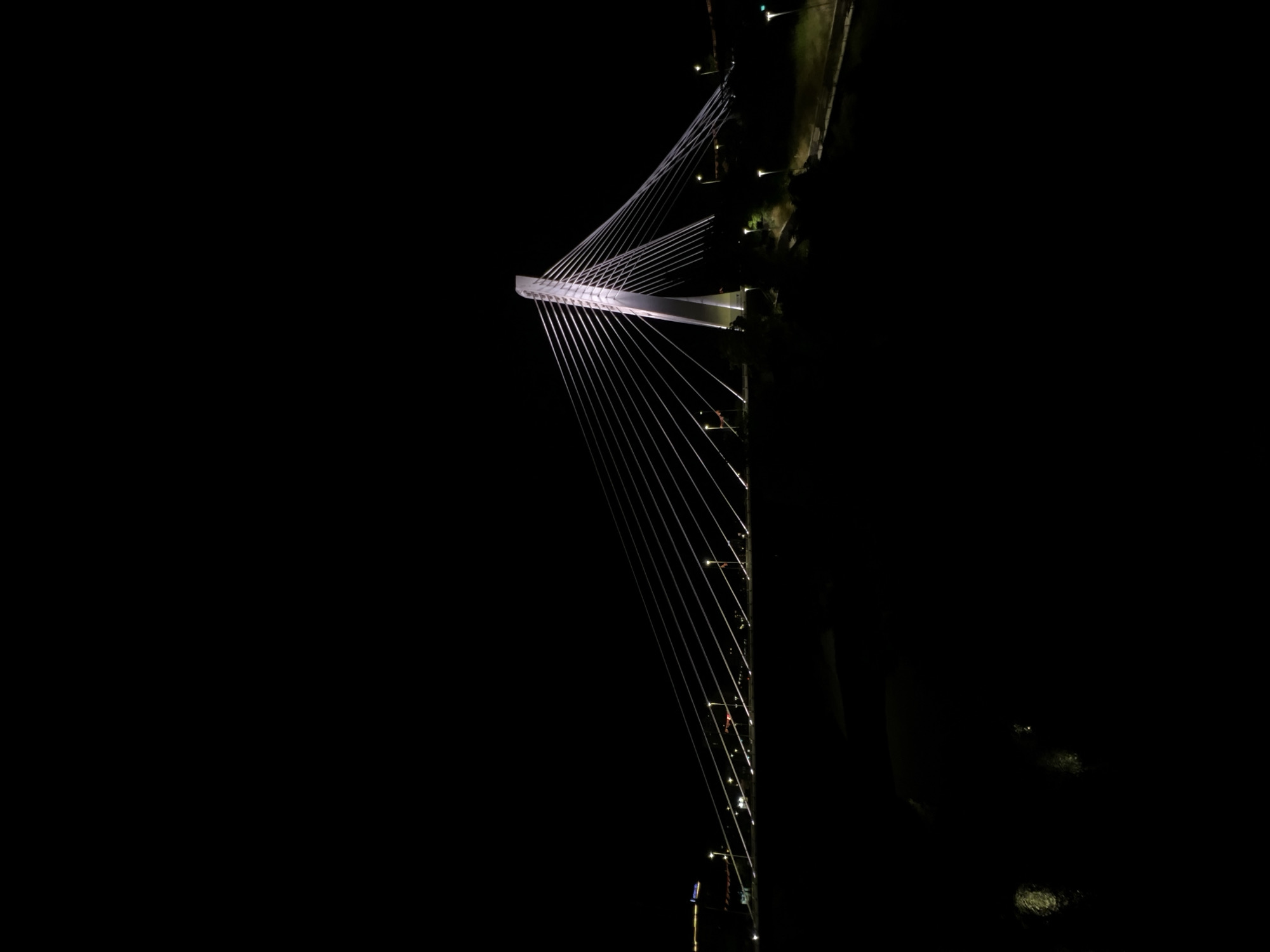 The image is a night-time, vertical shot of the Millennium Bridge in Podgorica, Montenegro. The bridge's iconic white pylon and its numerous suspension cables are illuminated, creating a striking silhouette against the dark sky. The cables fan out from the top of the pylon, supporting the roadway which is also subtly lit. Streetlights are visible along the edges of the bridge and on the surrounding landscape. The dark water of the Morača River is visible in the foreground, reflecting some of the ambient light. The scene is devoid of people. The overall impression is one of modern architecture illuminated against a natural, dark backdrop.