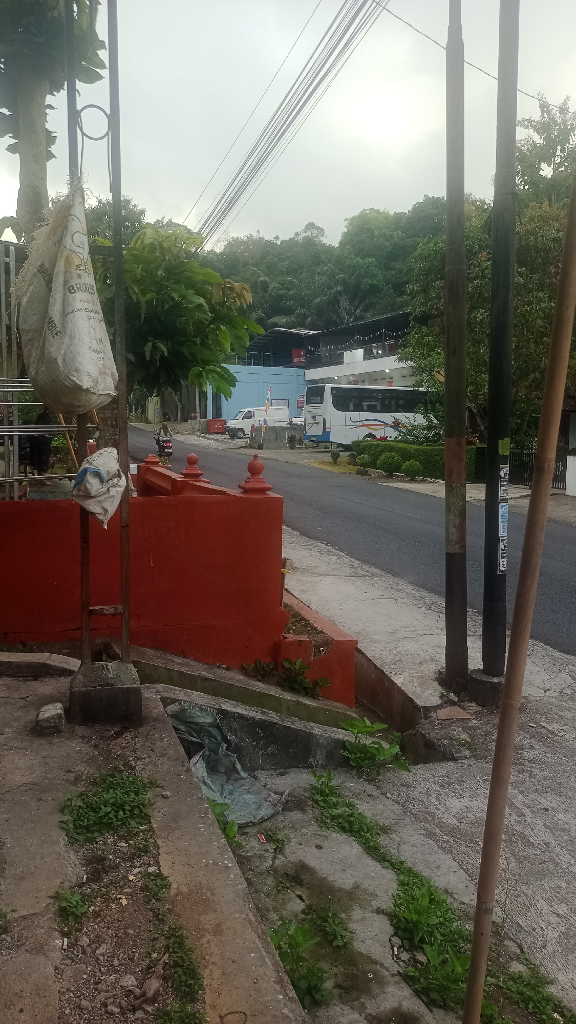 The image depicts a street scene in Karyamandala, Indonesia, under an overcast sky. In the foreground, a red-painted concrete barrier with decorative finials runs along the side of a road. A large, off-white woven sack, possibly containing animal feed, is hanging from a metal pole in front of the barrier. Below the barrier, a drainage ditch with some green foliage and debris is visible.
The road itself is paved and curves slightly to the right. On the opposite side of the road, a white bus with blue and yellow stripes is parked, alongside a white van. Behind the vehicles, a multi-story building with a dark roof and white facade is situated, bordered by lush green trees and vegetation that extends up a hillside in the background. Electrical wires stretch across the sky, supported by poles. The overall impression is of a semi-urban or semi-rural environment, possibly a commercial or transportation hub given the presence of the bus and building. No people are clearly identifiable or engaged in specific actions, though a motorcycle is seen in the distance on the road.