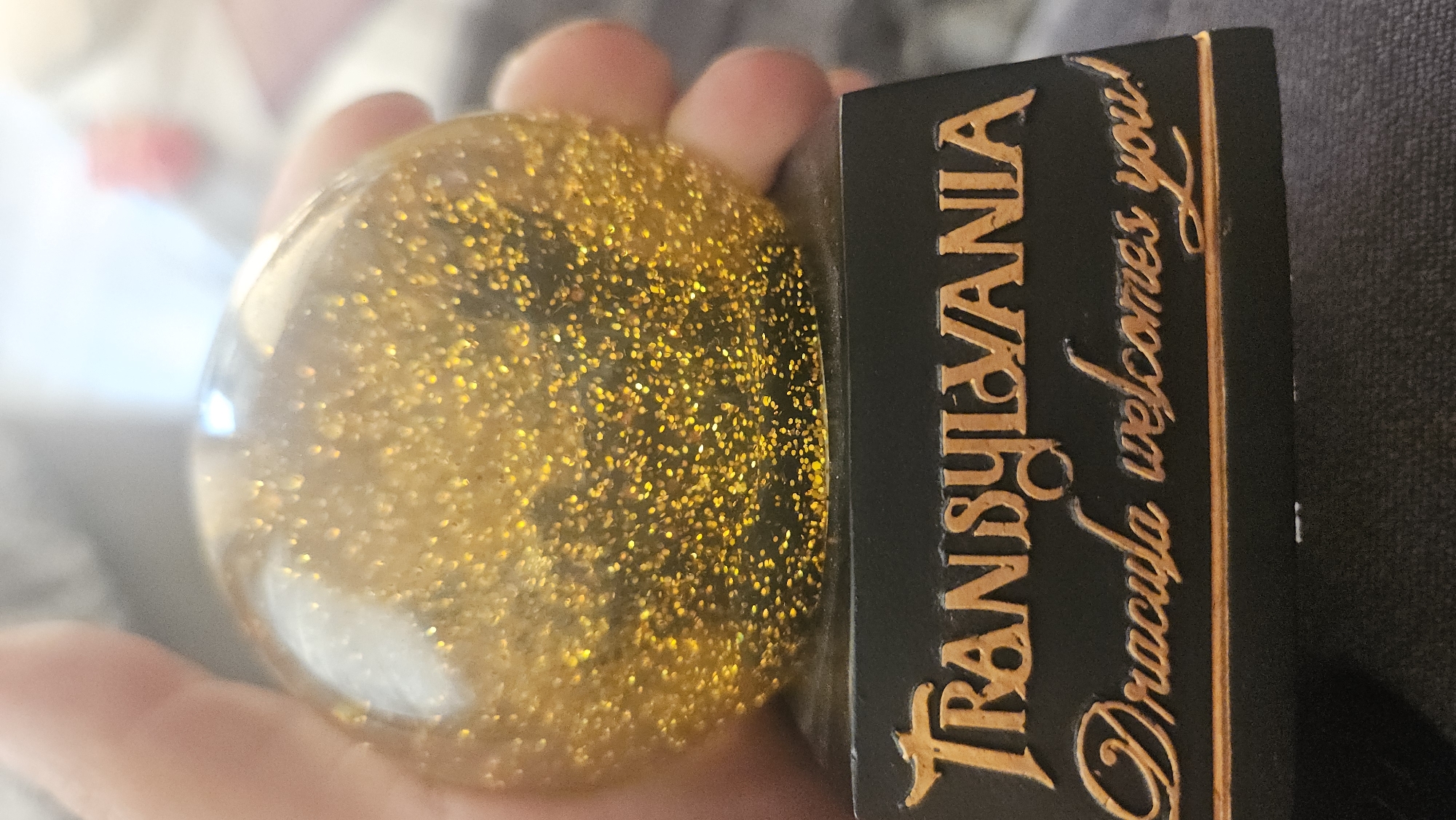 The image shows a hand holding a golden glitter-filled ball next to a black rectangular object. The object has gold-colored writing that reads "TRANSYLVANIA Dracula welcomes you!". The background is blurred and indistinct. The location is not discernible from the image.