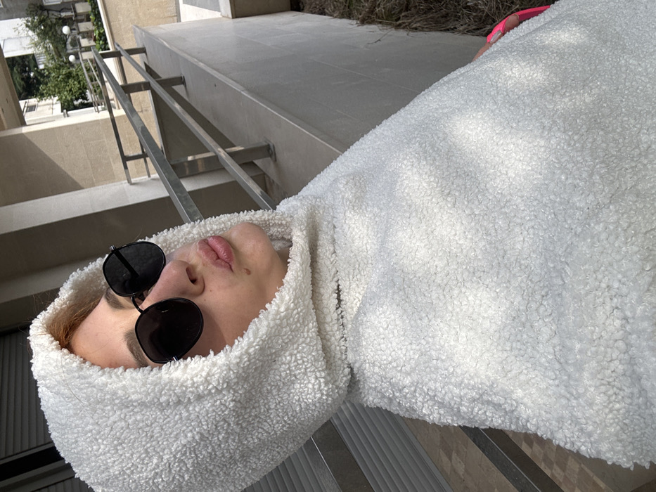 A person is depicted in a close-up, slightly upward-angled outdoor photograph within a residential area of Budva, Montenegro. The individual, likely female, occupies the majority of the frame, facing slightly right. They are wearing a white, textured sherpa or "teddy" fleece hooded garment with the hood drawn over the head, revealing light-colored hair at the hairline. Dark, hexagonal-shaped sunglasses with a thin dark frame obscure the eyes. The lips are pursed into a "kissing" or "duck face" expression, with a small mole visible on the upper lip. A glimpse of bright neon pink material is visible on the lower right edge of the frame, possibly part of an accessory or undergarment. The background, illuminated by direct sunlight creating strong shadows, features a light-colored concrete or stone wall and a multi-barred metallic railing extending diagonally. Behind these structures, a multi-story residential building with light-colored facades and balconies is discernible. Further back, green foliage from trees and shrubs, along with white globular outdoor light fixtures, suggests a landscaped urban environment.