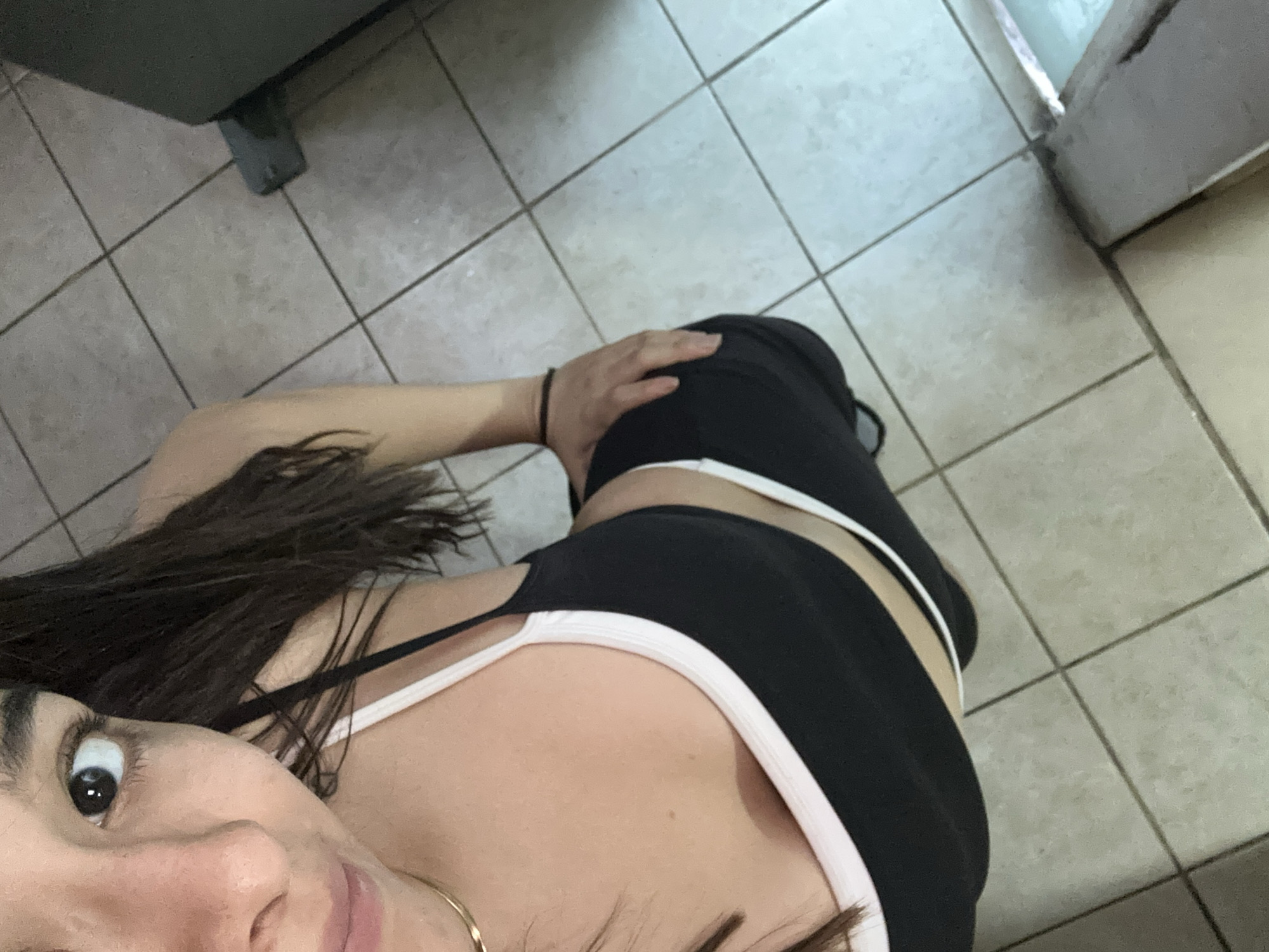 A woman is depicted in a first-person view selfie. She is wearing a black top with a white trim and black leggings. Her right hand rests on her thigh. She has long dark hair and is wearing a small gold hoop earring. The background consists of grey floor tiles. The location is most likely indoors. There is no information to determine that the location is in Puebla, Mexico.