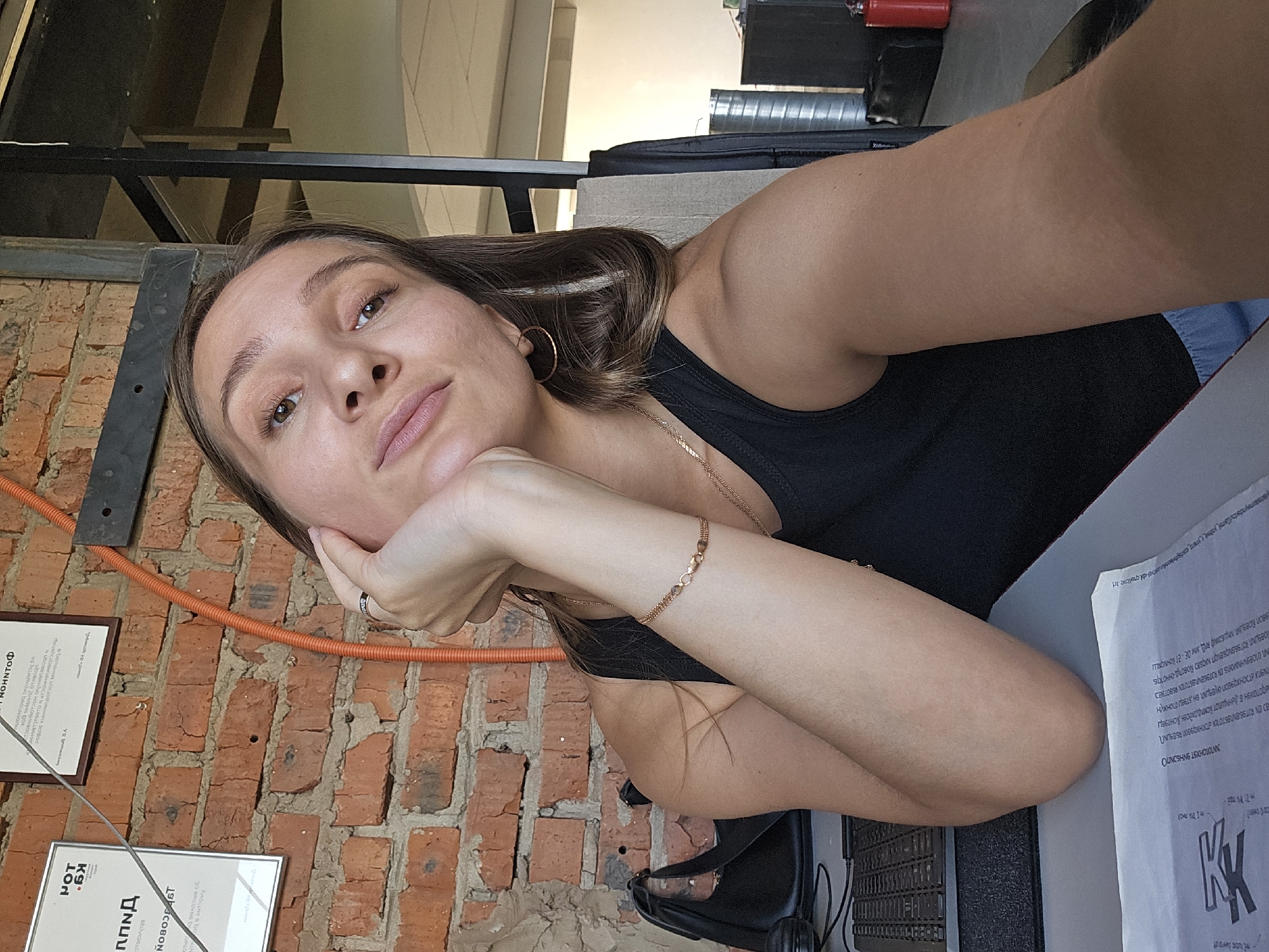 A young woman with brown hair and brown eyes is taking a selfie in what appears to be an office or workshop setting. She is wearing a black tank top and has a gold bracelet on her left wrist. Her left hand is propping up her chin, and she is looking directly at the camera. Behind her, a brick wall is visible, with a framed document hanging on it. To her left, a black handbag is placed on a desk next to a laptop. The environment seems to be indoors, with industrial-looking elements like metal beams and pipes. The specific location within the city of Chaikovsky, Russia, cannot be determined from the provided image.