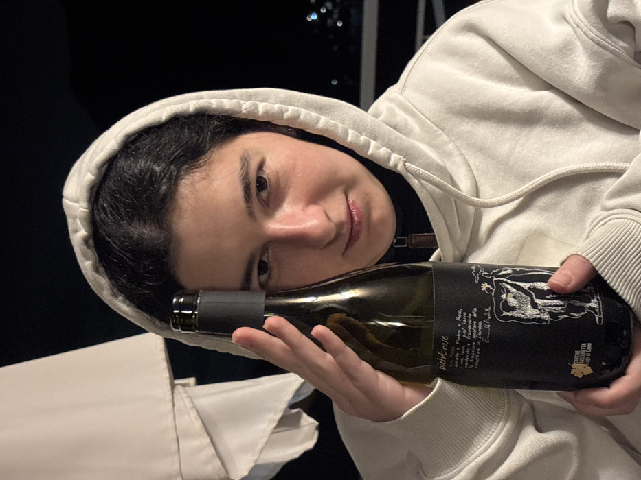 An individual is depicted holding a bottle of wine, captured in a medium close-up at night in Nesso, Italy. The subject is a young adult female with dark hair, wearing a light-colored hooded sweatshirt over a black collared top. She holds a dark green glass wine bottle with both hands, positioning it vertically such that the bottle's neck and upper body partially obscure her left eye and forehead. The bottle features a black label with white text, including "pietrErose" and "CANTINE ANGELINETTA Lago di Como", along with a stylized white drawing of two figures and a grape leaf logo. The subject looks directly at the camera with a neutral expression. In the background, a large white fabric structure, likely an awning or umbrella, is visible on the upper left. The distant background is dark, indicating nighttime, with faint light sources suggesting a view over a landscape.