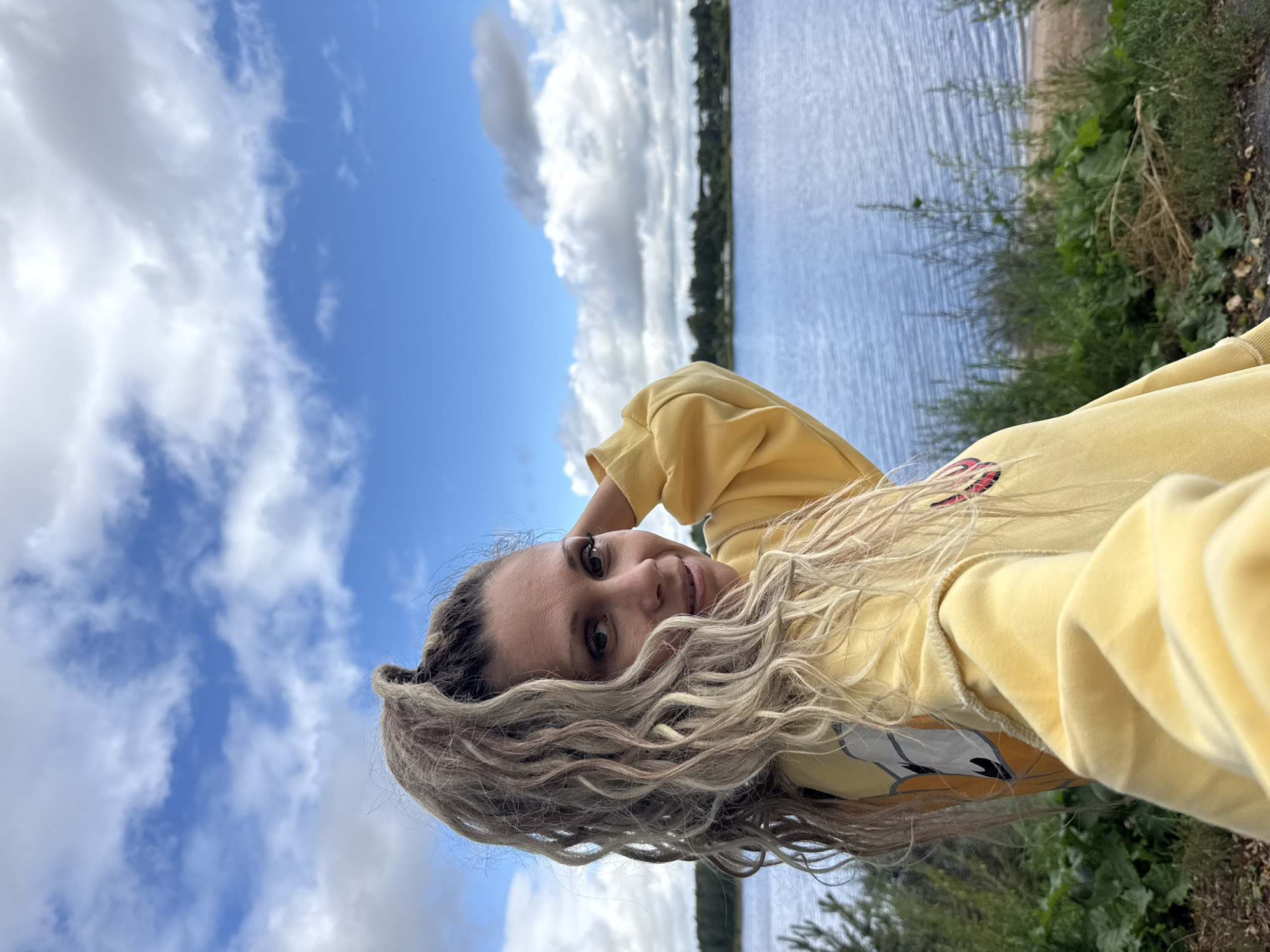 A woman with blonde dreadlocks tied into a high ponytail is posing for a selfie outdoors. She is wearing a yellow hooded sweatshirt with cartoon graphics on the front. She is looking at the camera and smiling, with her left hand resting on the back of her head. She is standing on a slight incline, with a body of water and a tree line in the background. The sky is blue with white clouds. The location is the city of Rusanovo, Russia.