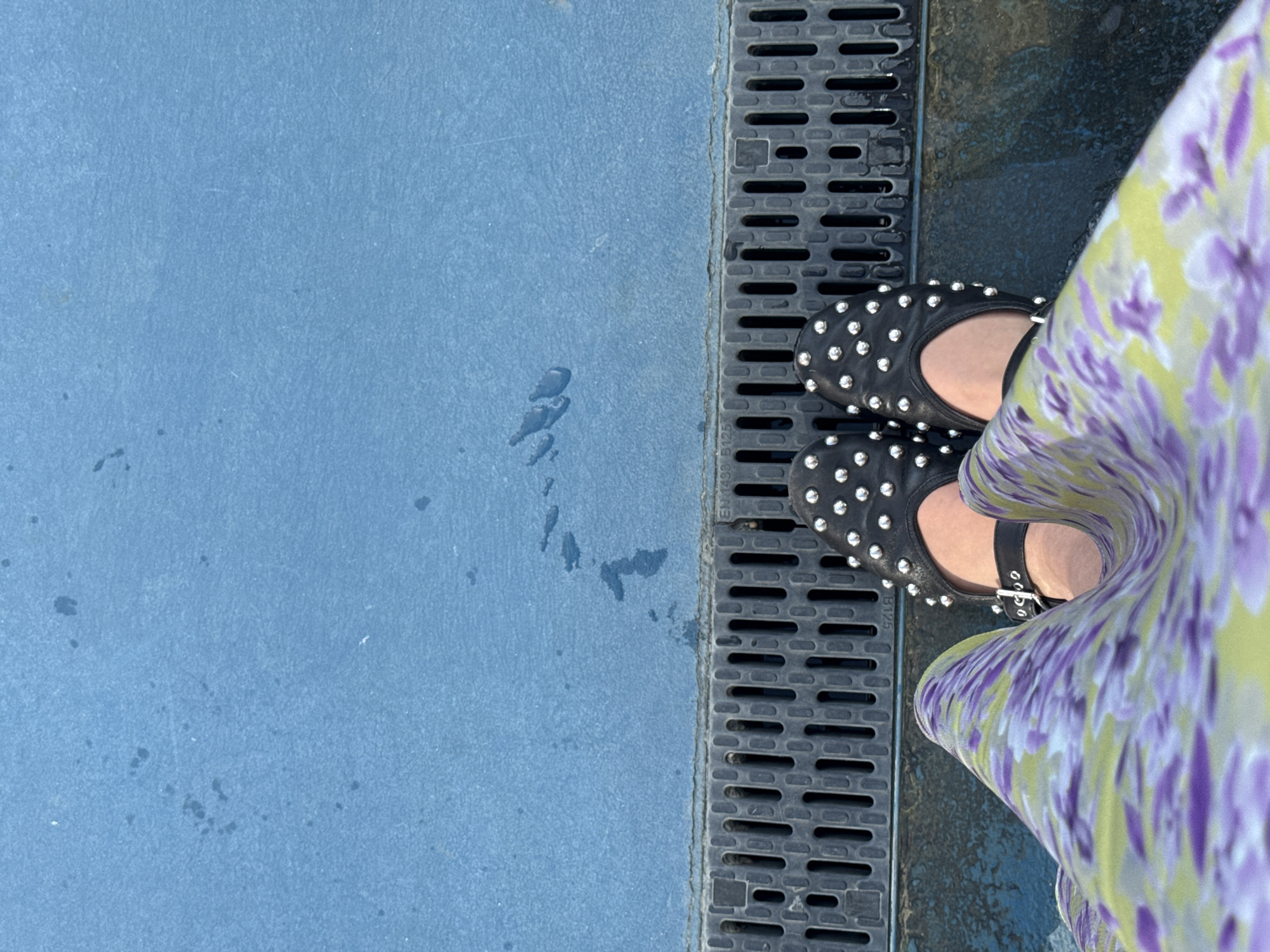 A top-down view captures the lower portion of a person's body standing over a drainage system on a blue surface. The person wears black, studded ballet flats or Mary Janes with a strap and buckle. A flowing dress or skirt, patterned with purple and lime green floral motifs, partially covers the legs. Directly beneath the feet is a dark gray or black linear drainage grate composed of rectangular slits. Markings "EN 1498" and "B125" are faintly visible on the drain cover, indicating a European standard for drainage systems. Beyond the drain, a broad expanse of textured, light blue ground is visible, upon which several distinct water droplets and splashes are scattered. The environment appears to be an outdoor public space, possibly a recreational area. The specific location in Milan, Italy, cannot be definitively identified from the provided visual information.