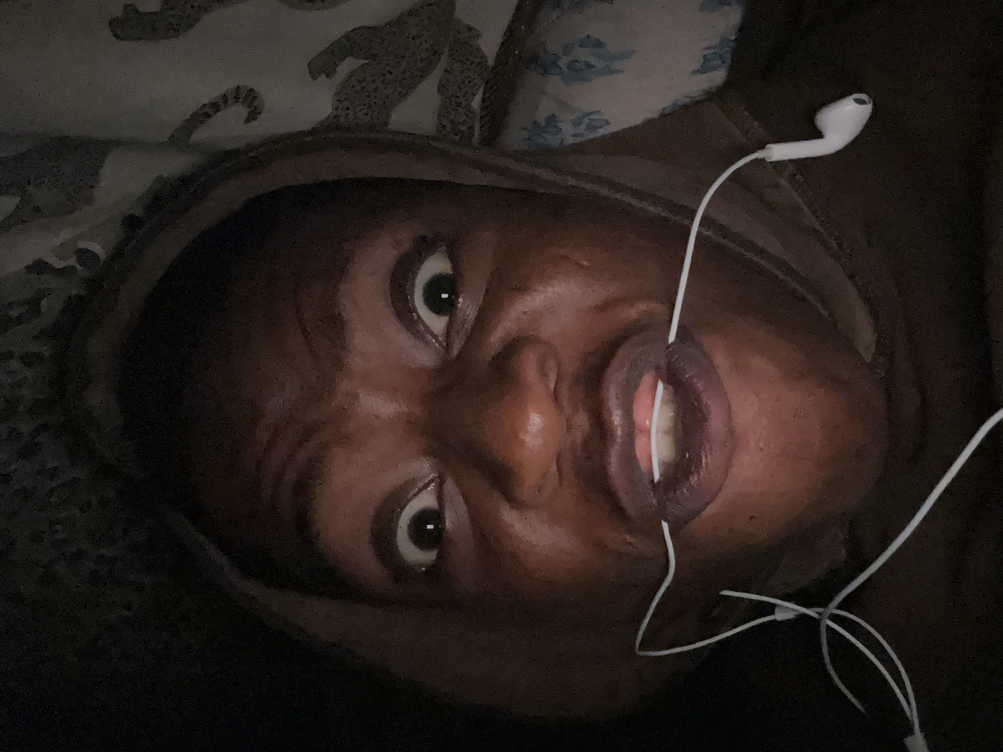 A close-up photograph depicts a person's face, wearing a brown hooded garment, and lying down with their head slightly tilted. White earbuds are connected to the subject's mouth. The background is blurry, revealing patterned bedding or fabric. The person's eyes are wide open, and their lips are slightly parted, showing their teeth. The setting appears to be indoors, possibly a bedroom. Location: Unknown, Unknown.