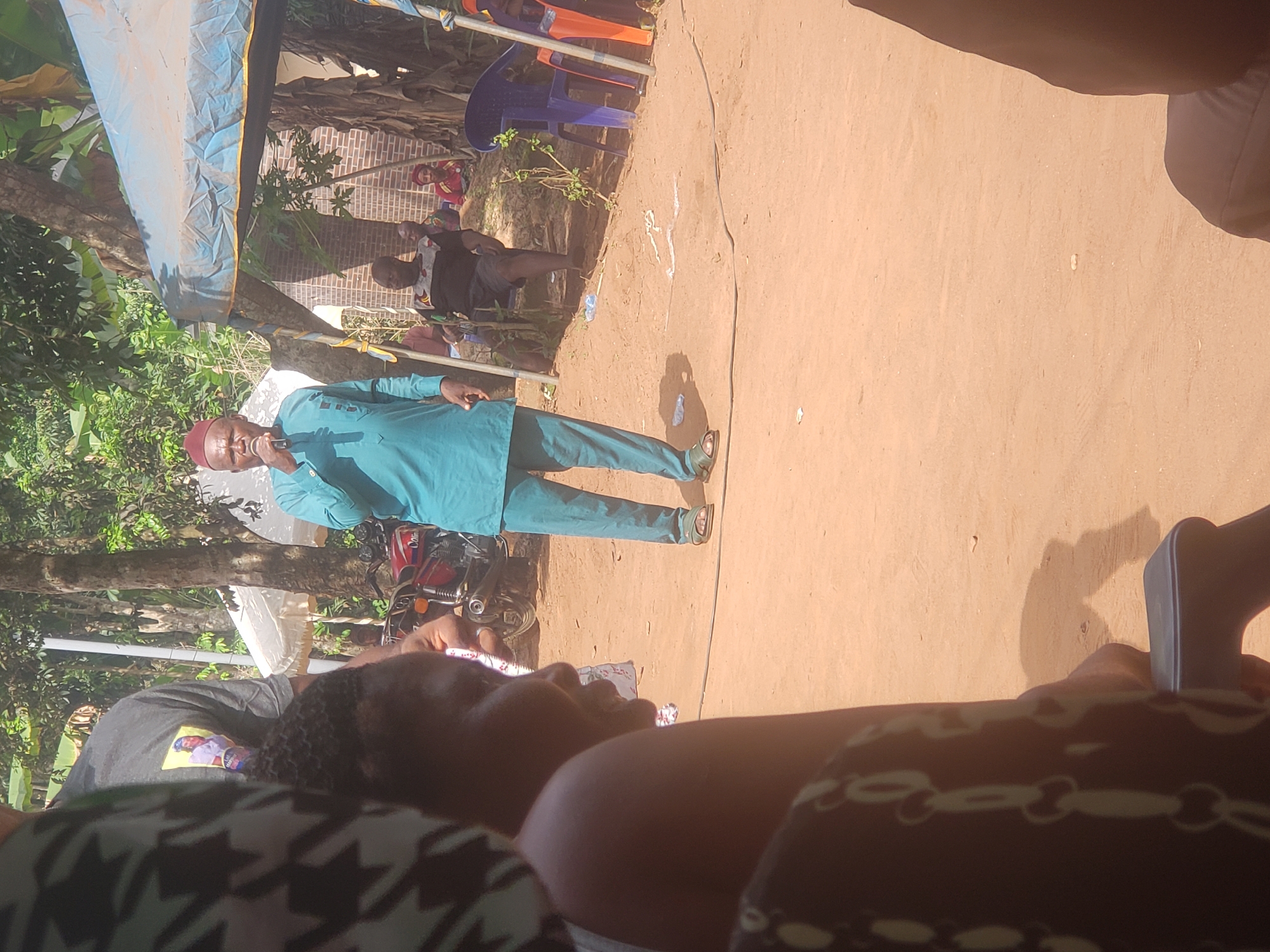 This outdoor image captures a community gathering or event under bright daylight in what appears to be a sunny, rural or semi-rural setting, consistent with Umundugba, Nigeria.
The central figure is a man standing on unpaved light brown earth, actively speaking into a microphone. He is dressed in a traditional long-sleeved teal tunic with matching trousers and a maroon cap. His attire suggests a formal or community leadership role.
An audience is present, with several individuals visible in the foreground and background. In the immediate foreground, the back of a person's head and shoulder are seen, adorned with braided hair and wearing a dark, possibly patterned, garment. Another person's leg and foot are partially visible in the extreme lower right.
In the background, on the right side, several people are seated on blue and orange plastic chairs. One person behind them wears a black t-shirt with an indecipherable graphic print. Further back, a red brick wall is visible. To the left and behind the speaker, a red motorcycle is parked next to a tree. Lush green trees and foliage frame the left side of the scene.
A light blue tarp or canopy stretches across the upper left portion of the image, providing shade. The strong shadows cast by the subjects and objects indicate a bright, sunny day. The event appears to be a public address or a community meeting, with the speaker engaging an assembled audience. No discernible text is clearly visible on clothing or objects, other than a pattern on the speaker's shirt and an unreadable graphic on a background figure's shirt.