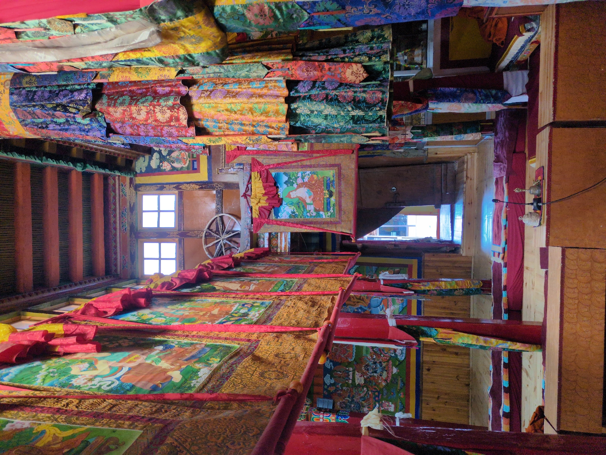 This image shows the interior of a room within a building, likely a monastery, in Yulang, India. The room is narrow and long, with high ceilings supported by wooden beams. The walls are adorned with colorful thangka paintings and intricate fabric hangings, predominantly in red, yellow, blue, and green hues, featuring floral and religious motifs. Red wooden pillars line the sides of the room, supporting horizontal beams from which more fabric decorations hang. In the distance, a doorway leads to another section of the building, and two square windows are visible high on the wall, allowing natural light to enter. The floor is made of wooden planks, and red mats are laid out, suggesting a space for meditation or prayer. In the foreground, low wooden tables are present, and on one of these tables, a microphone and some bells are visible, indicating the presence of sound equipment and possibly a ceremonial setup. The overall atmosphere is serene and rich with religious and cultural artifacts. There are no people visible in the image.