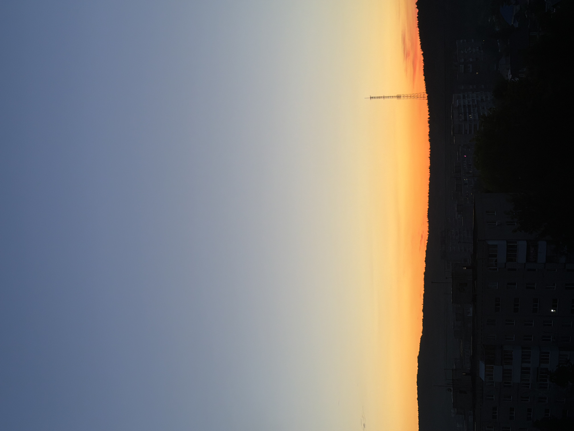 The image depicts an urban landscape at dusk or dawn. The sky transitions from blue to orange, suggesting the sun is either rising or setting. A tall transmission tower is visible on the horizon against the orange sky. The silhouette of a city skyline with apartment buildings and trees is situated below the sky. Buildings appear to be in a state of dim light. The image orientation is rotated.