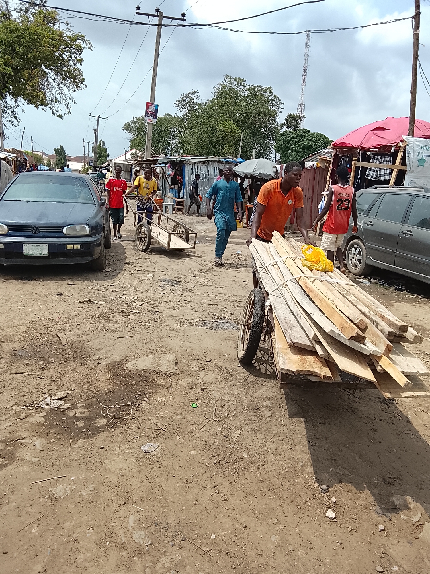 The image depicts a street scene in Abuja, Nigeria. The ground is bare dirt with scattered debris. Four men are present, one in an orange shirt pushing a two-wheeled cart loaded with lumber, another in a blue garment walking nearby, and two more, one in a red shirt and one in a yellow shirt, pulling an empty cart. Two cars are visible: a dark blue VW Golf on the left and a gray station wagon on the right. Background elements include ramshackle stalls, a utility pole, a telecommunications tower, and other people.