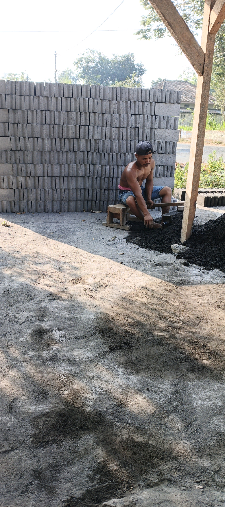 A shirtless man is sitting on a wooden stool in front of a large stack of gray cinder blocks. He is wearing blue shorts and a dark baseball cap. The man is holding a wooden tool, possibly a trowel or shovel, and appears to be working with a pile of dark, granular material in front of him, which could be cement or dirt. The scene is outdoors, with a dirt and concrete ground. In the background, there is more of the cinder block wall, some vegetation, a wooden support structure, and a house with a tiled roof. The lighting suggests it is daytime, with sunlight casting shadows. The image seems to be taken in Kasiyan, Indonesia, a location known for its brick and cinder block production.