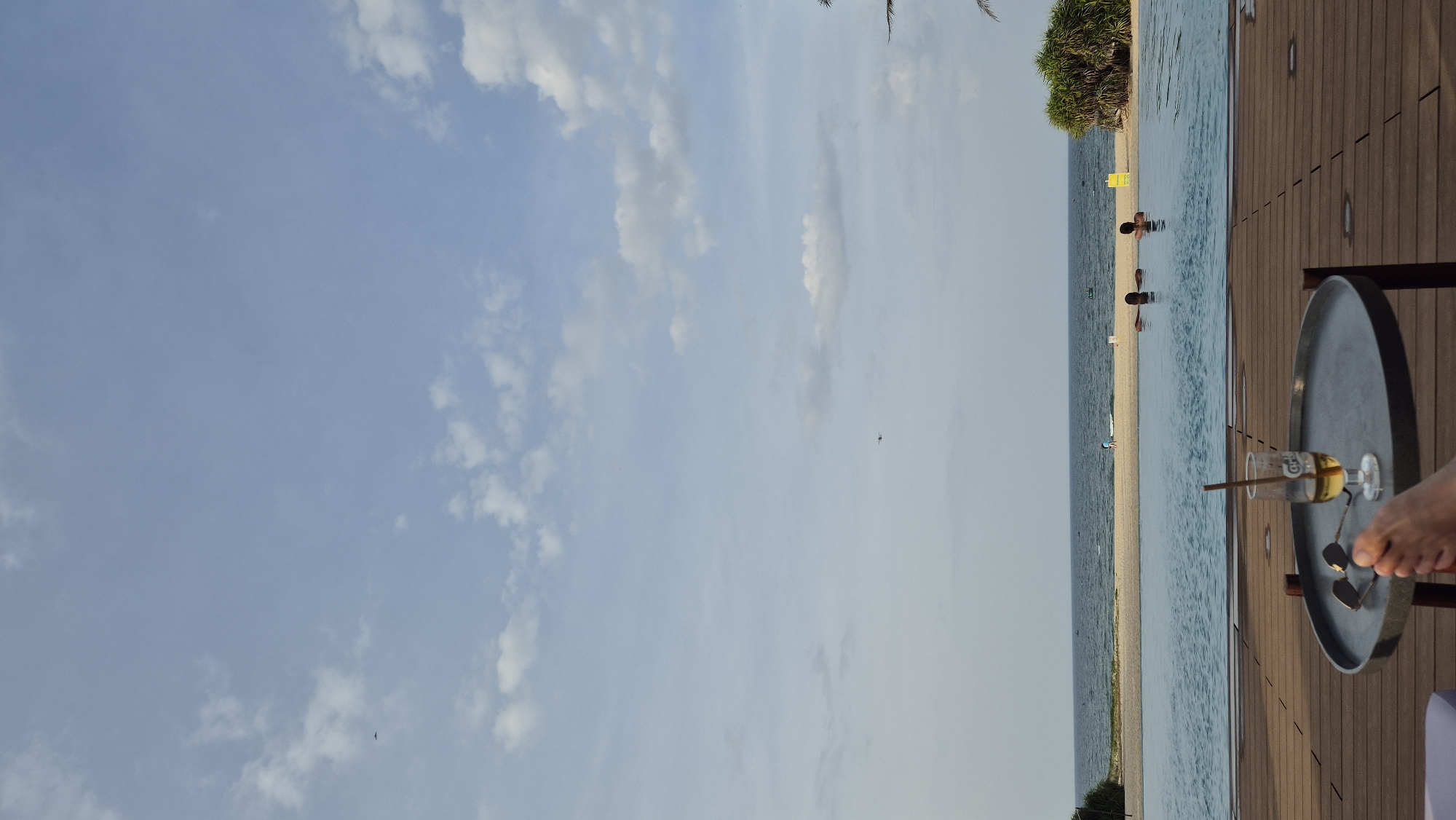 In Hoi An, Vietnam, a scene unfolds by the sea. The foreground features a wooden deck with a circular coffee table, upon which rests a glass with a light-colored beverage and a straw, accompanied by a pair of sunglasses. A person's foot is visible in the lower left corner. Beyond the deck lies an infinity pool filled with blue water, where two individuals are partially submerged. In the midground, a sandy beach stretches out, with the ocean meeting the horizon under a sky dotted with clouds. A small boat is visible on the water, and two figures can be seen on the beach. Lush green foliage borders the beach on the right side, and a palm tree is partially visible. The sky is a pale blue with scattered white clouds, and two distant dark specks, possibly birds or small aircraft, are present in the sky.