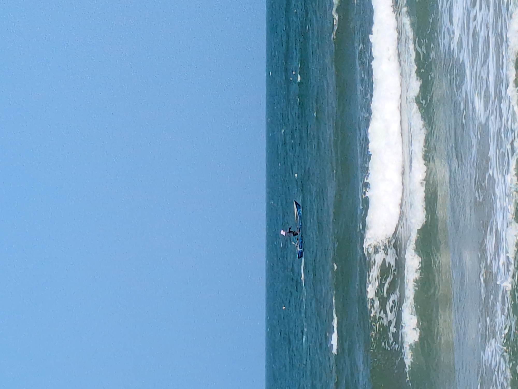 The image shows a partially obscured, rotated view of the ocean and sky. On the left, a clear blue sky is visible. On the right, the ocean is present with visible waves and white foam. In the middle of the water, a person appears to be windsurfing. The location may be South Quy Nhon Ward, Vietnam.