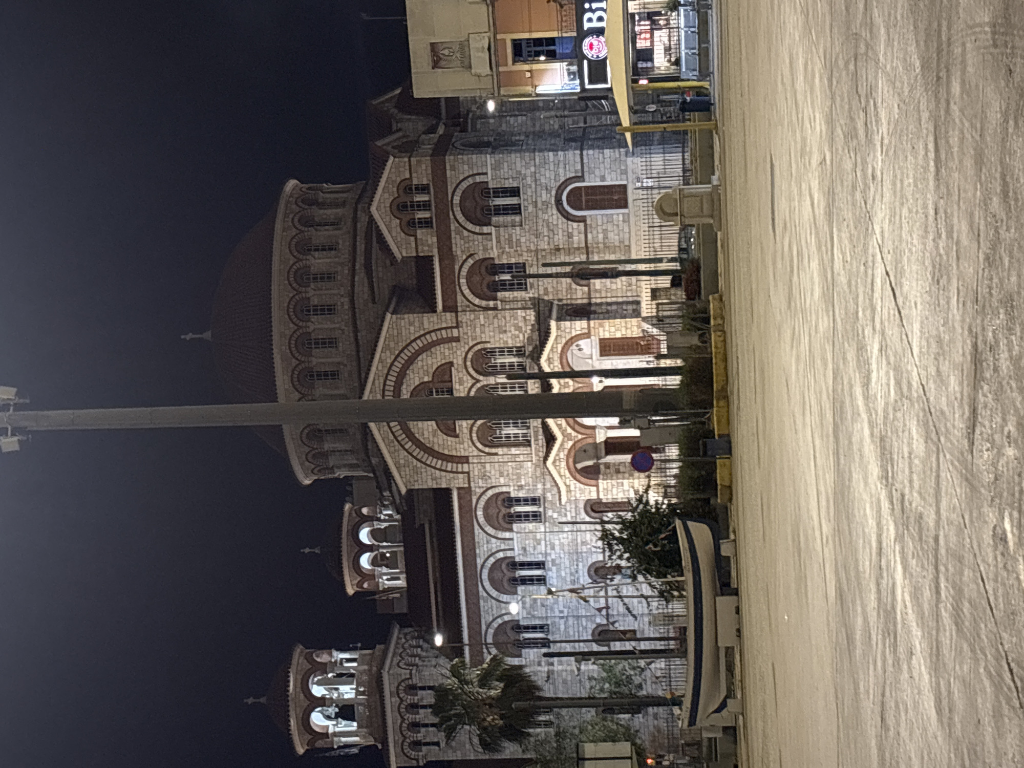 The image shows the Church of Saint Nicholas in Piraeus, Greece at night. The Byzantine-style architecture of the church is visible, including its domes, arches, and intricate stonework. Streetlights illuminate the building's facade. Adjacent to the church are other buildings, including one with a visible "Bi" sign. The scene includes the pavement in front of the church, with the presence of a boat. The sky is dark, indicating the night setting.