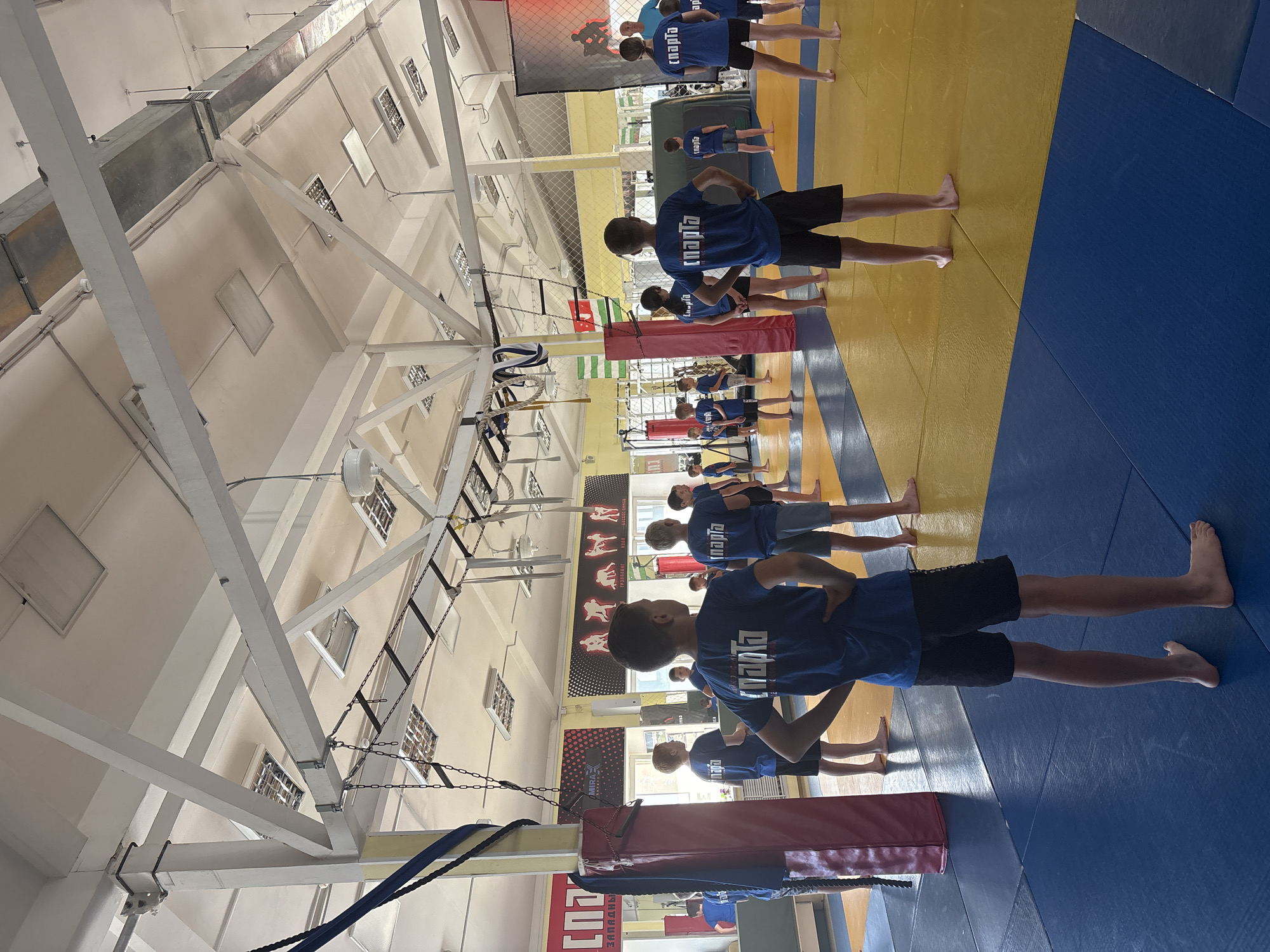 In a bright, indoor gymnasium with high ceilings and exposed beams, a group of young people, appearing to be mostly boys, are lined up on a blue and yellow mat. They are all wearing matching blue t-shirts with white lettering and black shorts. The t-shirts have the word "СПОРТА" (SPORTA) printed on them. The participants are standing in a row, facing away from the camera, and seem to be engaged in a martial arts or athletic training session. Some of the individuals have their hands on their hips, suggesting they are awaiting instructions or in a static pose.
The setting includes gym equipment such as ropes and padded mats. Large posters depicting various wrestling or judo poses are visible on the wall in the background, further indicating the nature of the activity. The lighting appears to be artificial, suggesting it is daytime or the gym is self-lit. There is no visible indication of weather conditions. The overall atmosphere seems focused and disciplined, characteristic of a training environment. The location is identified as Chaikovsky, Russia.