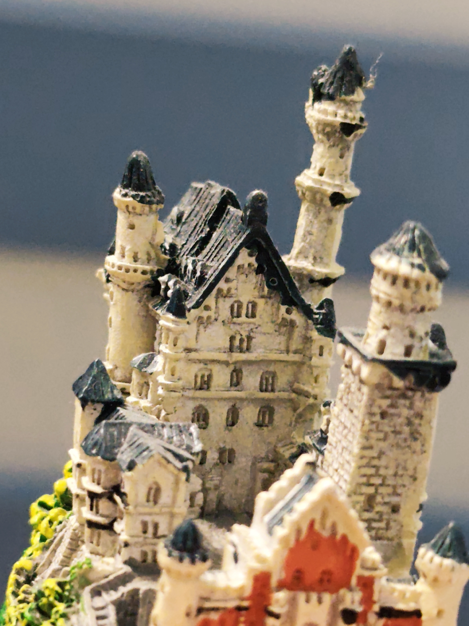 This image presents a detailed, close-up view of a miniature model castle, likely representative of Neuschwanstein Castle, set against a soft, out-of-focus blue-grey background. The model features intricate architectural details, including multiple turrets, towers, and sections of the main castle structure. The primary color of the castle walls is an aged cream or off-white, with prominent dark grey or black roofs on various spires and main building sections. Subtle green foliage elements are visible at the lower left base, and a distinct red-orange roof section is present on a lower building towards the right. Textured surfaces mimic stone or brickwork, and numerous small, dark openings represent windows or decorative elements. A thin, light-colored fiber is visibly attached to the peak of the tallest, slender spire on the upper right side of the model. The shallow depth of field ensures the focus is entirely on the miniature, blurring the background into a uniform, indistinct tone. The scene is situated in the city of Pazardzhik, Bulgaria.