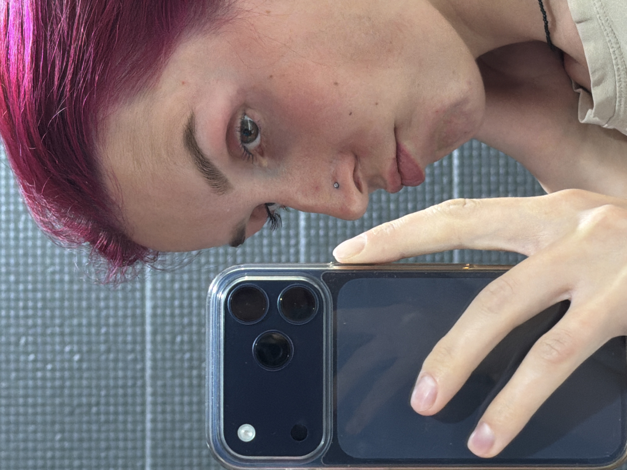A close-up, low-angle selfie shows a person holding a dark-colored smartphone with a multi-lens camera system in their left hand. The person's face is angled towards the camera, with their right side more visible. They have bright pink hair, a nose piercing, and are wearing a light-colored top with a dark beaded necklace. The background appears to be a tiled wall. The image does not contain any information that would allow for the identification of the city of Chaikovsky, Russia.