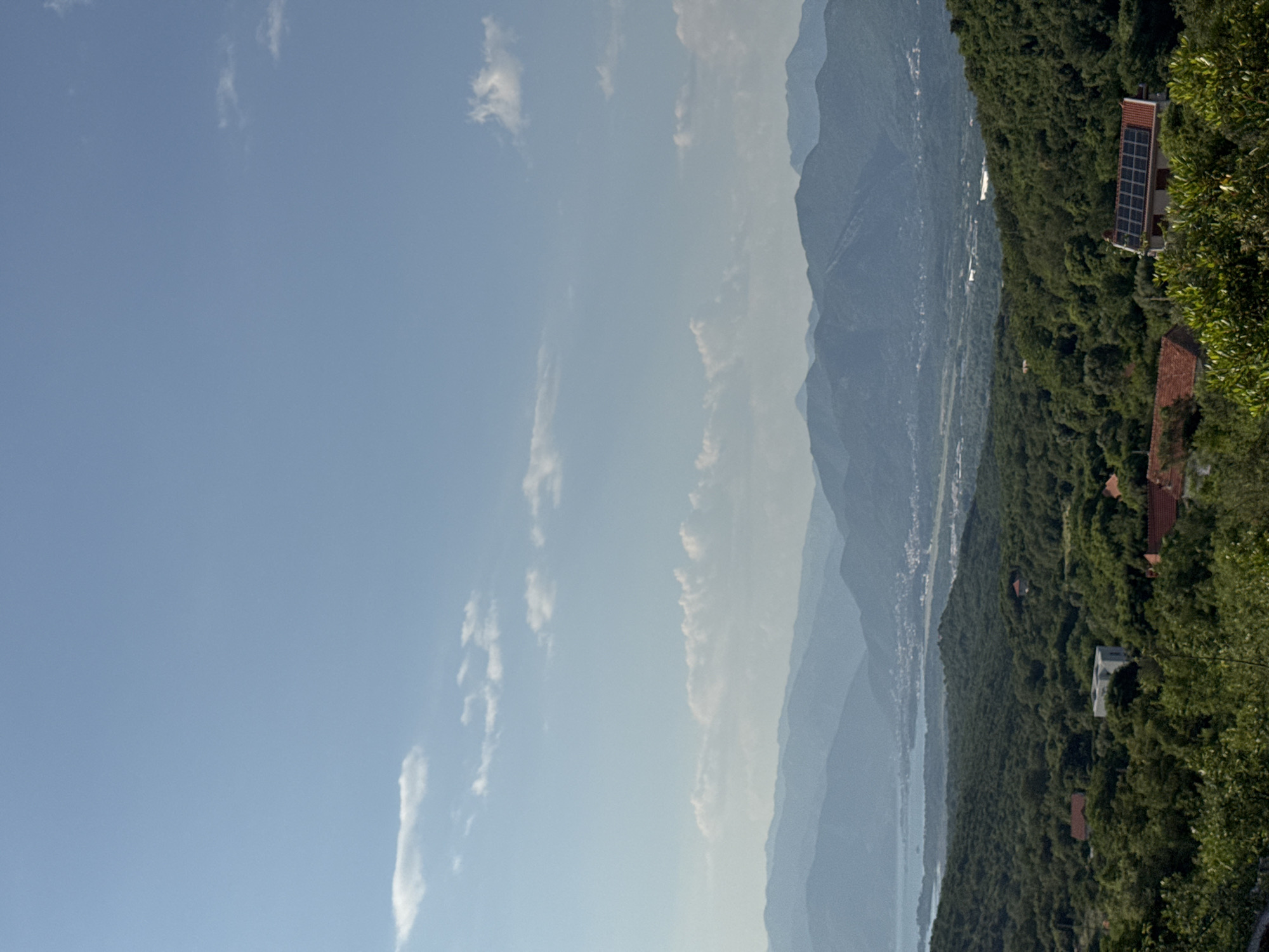The image shows a scenic landscape with a view of mountains and a body of water, likely in Kubasi, Montenegro. The sky is a pale blue with scattered white clouds. The mountains are layered, fading into the distance with a blueish hue. Lush green vegetation covers the slopes closer to the viewer. Several buildings with terracotta roofs are nestled among the trees, and one building has solar panels visible on its roof. The image is taken from an elevated position, providing a wide view of the landscape.