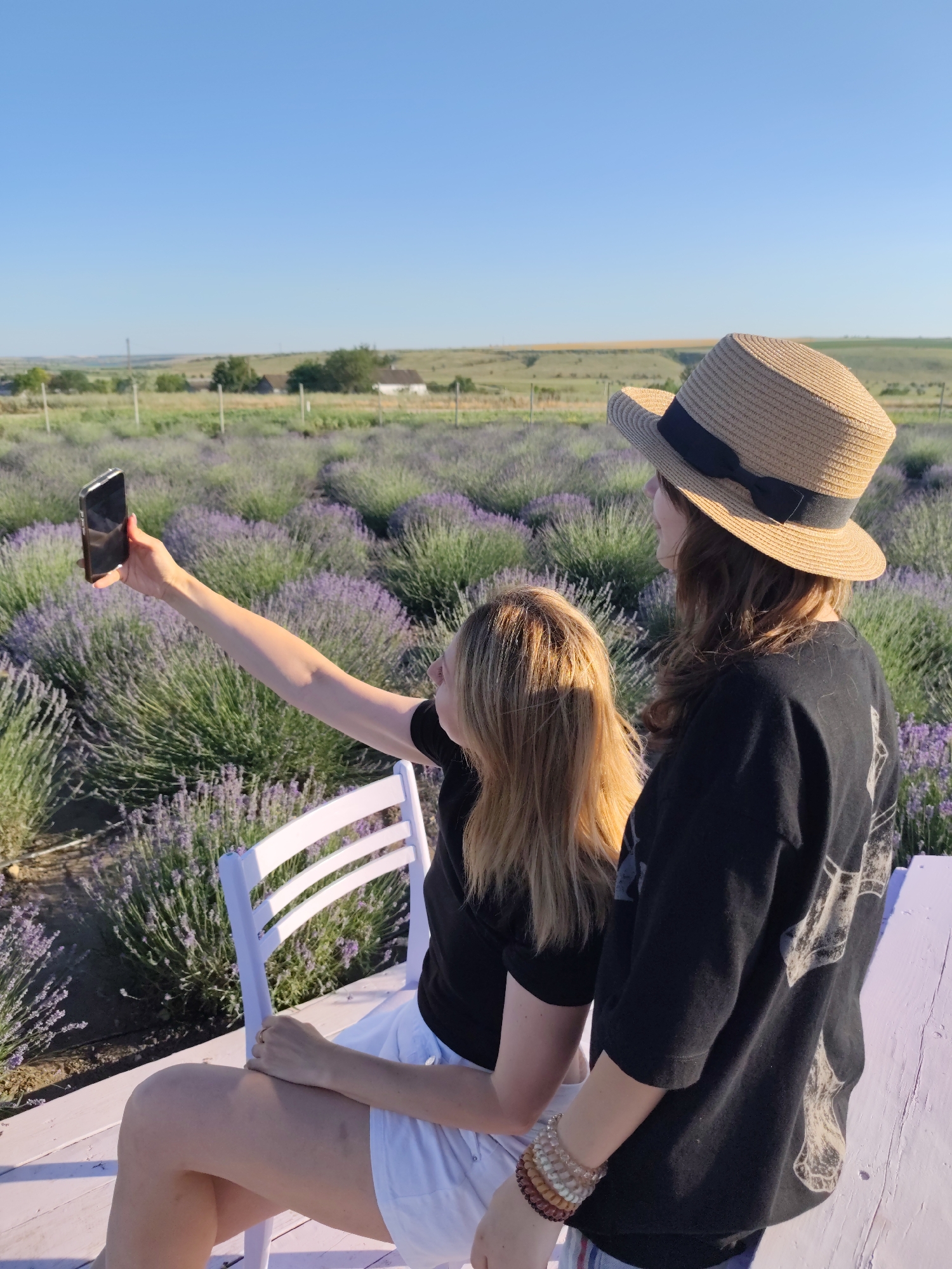 In Nechaiane, Ukraine, two women are taking a selfie in a lavender field under a clear blue sky, suggesting a pleasant day. The woman on the left, sitting on a white chair, holds up a smartphone to capture the moment. She has long blonde hair and is wearing a black t-shirt and white shorts. The woman on the right, standing, wears a straw hat with a black ribbon and a black t-shirt. She is adorned with several beaded bracelets.
The expansive lavender field, with its vibrant purple hues, stretches out behind them, bordered by distant green fields and a few scattered buildings under the bright, sunny sky. The lighting indicates it is either early morning or late afternoon, casting a warm glow on the scene. The overall atmosphere appears relaxed and cheerful, as the two women enjoy their time together in the picturesque lavender field. No visible text is present in the image.