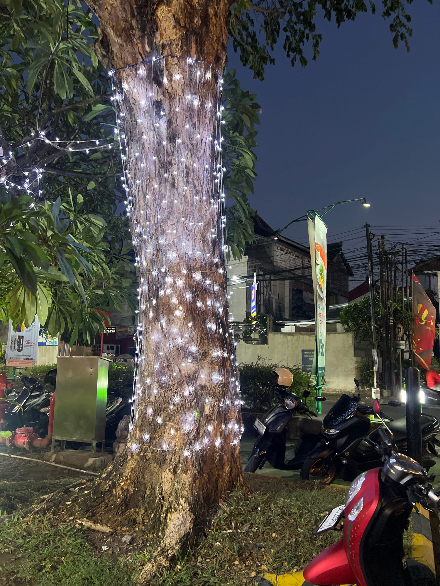 The image depicts a street scene in Kuta, Indonesia, at dusk or early evening. A large tree trunk is prominently featured in the foreground, wrapped with numerous white string lights that illuminate its textured bark. To the left of the tree, lush green foliage is visible. Beyond the tree, a row of parked motorcycles, mostly scooters, lines the street. Some of these motorcycles are dark-colored, while one in the immediate foreground is red. A street lamp with a curved arm extends overhead, supporting two vertical banners. One banner is predominantly white with an image of food and text, while another banner further down the street is dark red with yellow accents. Buildings are visible in the background, appearing to be of various architectural styles. Utility poles and wires are also present, indicating a developed urban environment. The ground around the tree is covered in grass and a paved area where the motorcycles are parked. The overall atmosphere is that of a moderately busy street in Kuta, likely near a commercial or entertainment district, with decorative lighting adding a festive element.