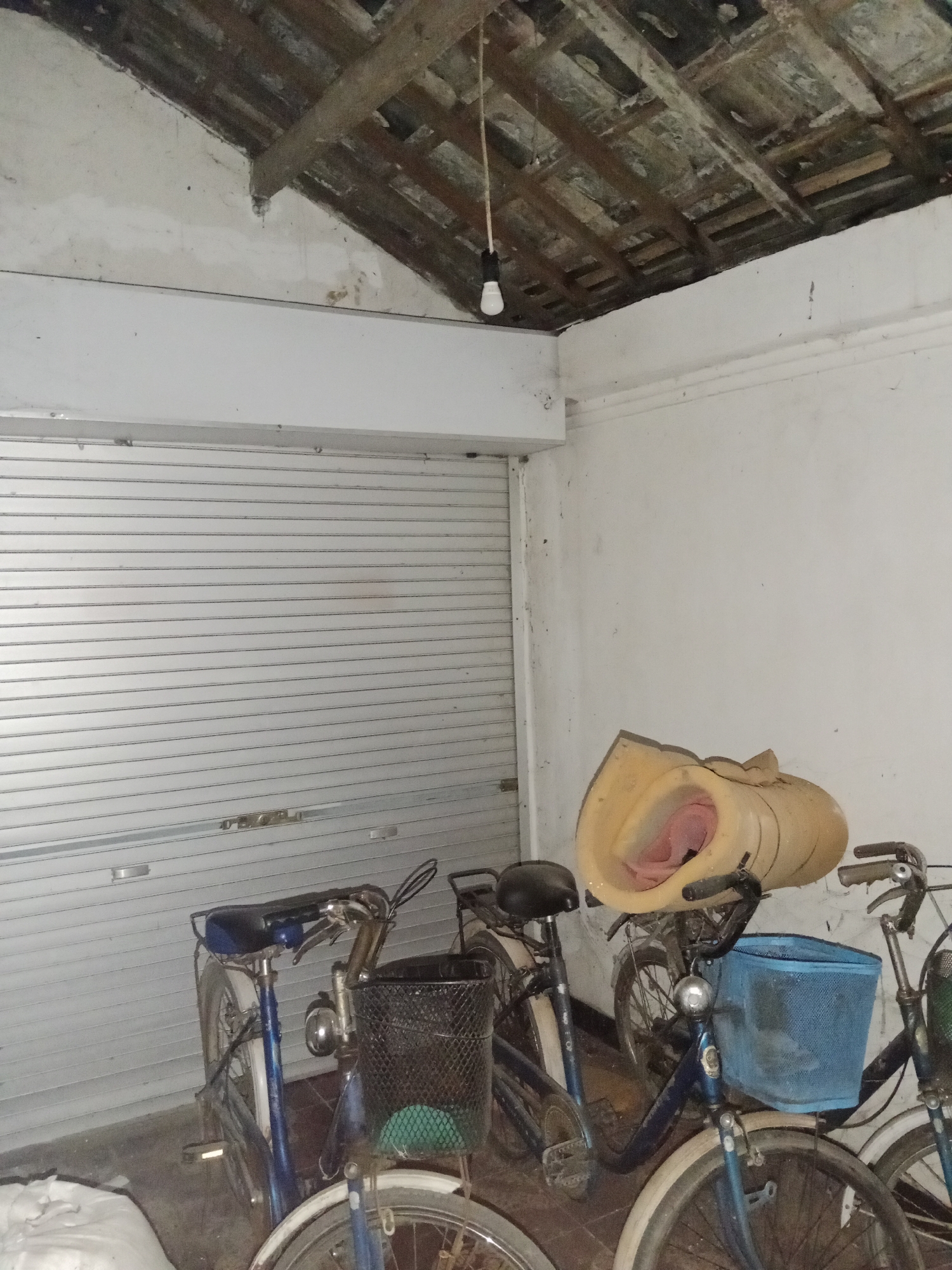 In a dimly lit, enclosed space, likely a garage or storage area, multiple bicycles are parked against a metallic, roll-up door. The ceiling is exposed, revealing wooden beams and rafters, with a single light bulb hanging from a wire. To the right, a large, yellow, foam-like object with a pink interior rests on top of a bicycle. The floor appears to be concrete. The scene is located in Ringinrejo, Indonesia, indicated by the surrounding environment.