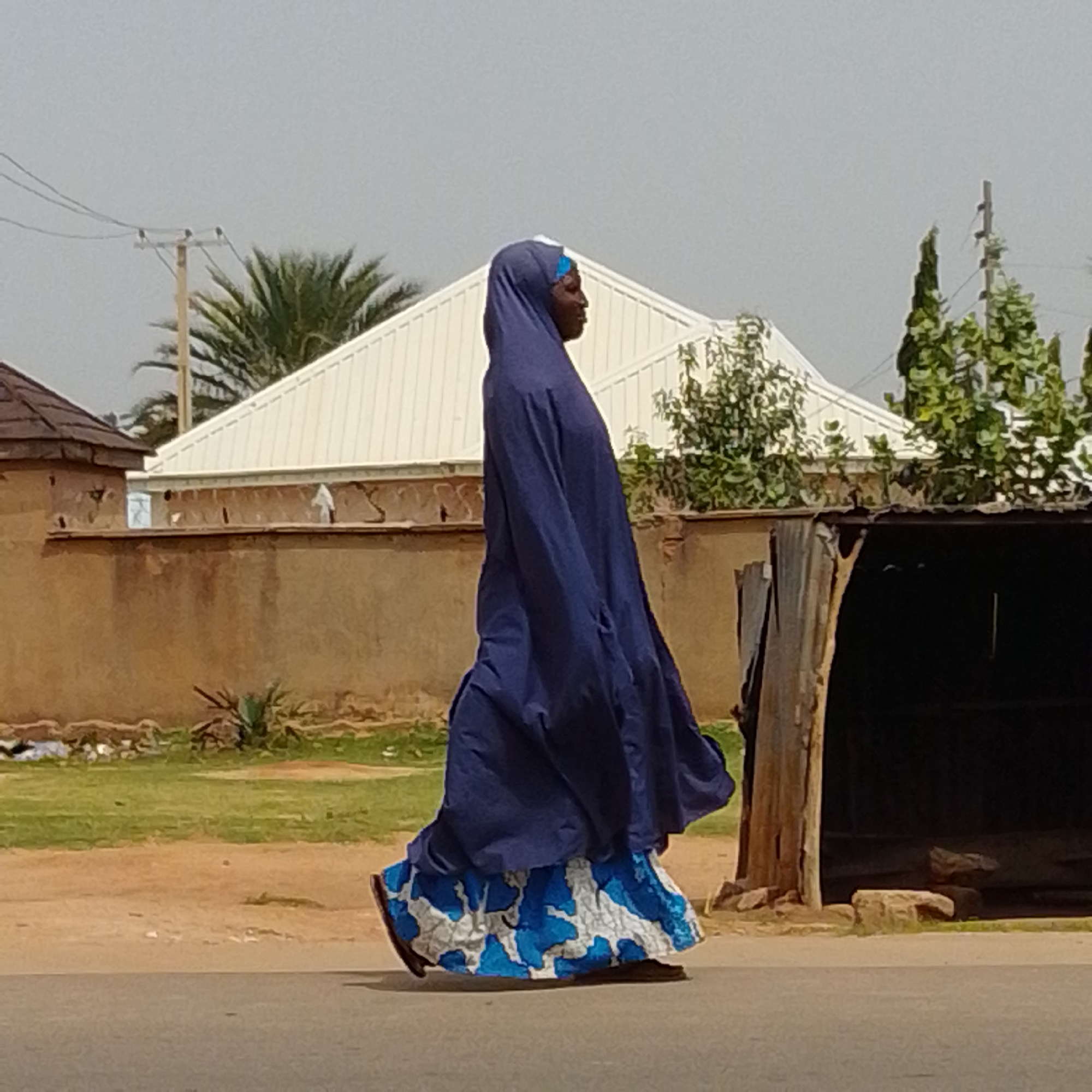 A woman in a long navy blue robe and a blue headscarf walks from left to right across a paved road in Gombe, Nigeria. She is also wearing a white and blue patterned skirt. Behind her, a tan-colored wall runs horizontally across the image, with a light-colored corrugated metal roof structure partially visible to the right. In the background, there are palm trees and other green foliage, with a white, gabled building and a utility pole with wires. The sky is overcast and a pale gray.