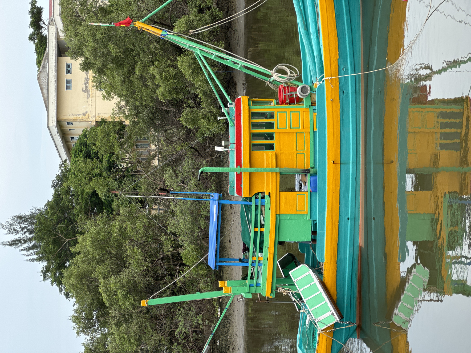 A brightly colored fishing boat is docked in a calm body of water, with its reflection clearly visible. The boat has a turquoise hull with a yellow stripe along the upper edge. The superstructure is a vibrant yellow with a red roof. Green accents are visible on the railings, masts, and other structural elements. A small blue canopy is attached to the cabin. The boat is equipped with fishing gear, including a large green net attachment. In the background, lush green trees and vegetation line the water's edge. Behind the trees, a multi-story building with a pale yellow facade and a flat roof is partially visible. The sky is overcast. The scene is likely located in the coastal region of Phú Thủy, Vietnam, a place known for its fishing communities and proximity to natural waterways. There are no people clearly visible on the boat or in the immediate vicinity, though it is implied to be in active use. The overall impression is one of a tranquil, working harbor environment with a focus on maritime activity.