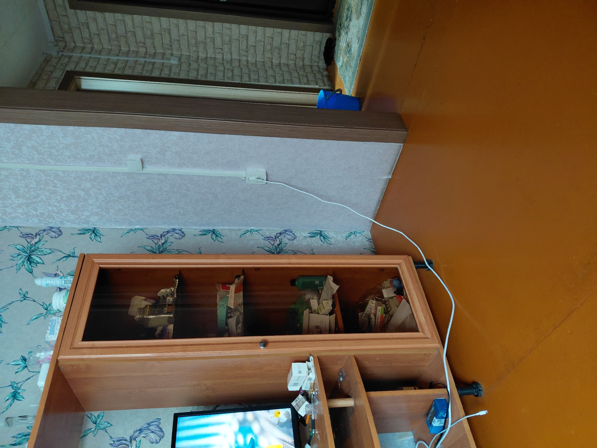 This is a medium-angle, full shot of an indoor scene, likely a room in a residential apartment in Malinovka, Russia. The dominant features are a light-wood entertainment unit with a glass-doored cabinet filled with various items, and a wall with a light-colored floral wallpaper. A television screen is visible on the left side of the entertainment unit. A white electrical cord runs from a power outlet on the wall, across the floor, and connects to something on the entertainment unit. To the right of the entertainment unit, a doorway leads to another room with brick-patterned wallpaper. A blue bucket is visible in the adjacent room. The floor is a solid, orange-brown color. The lighting suggests it is daytime, with light coming from an unseen window. There are no people visible in the image.