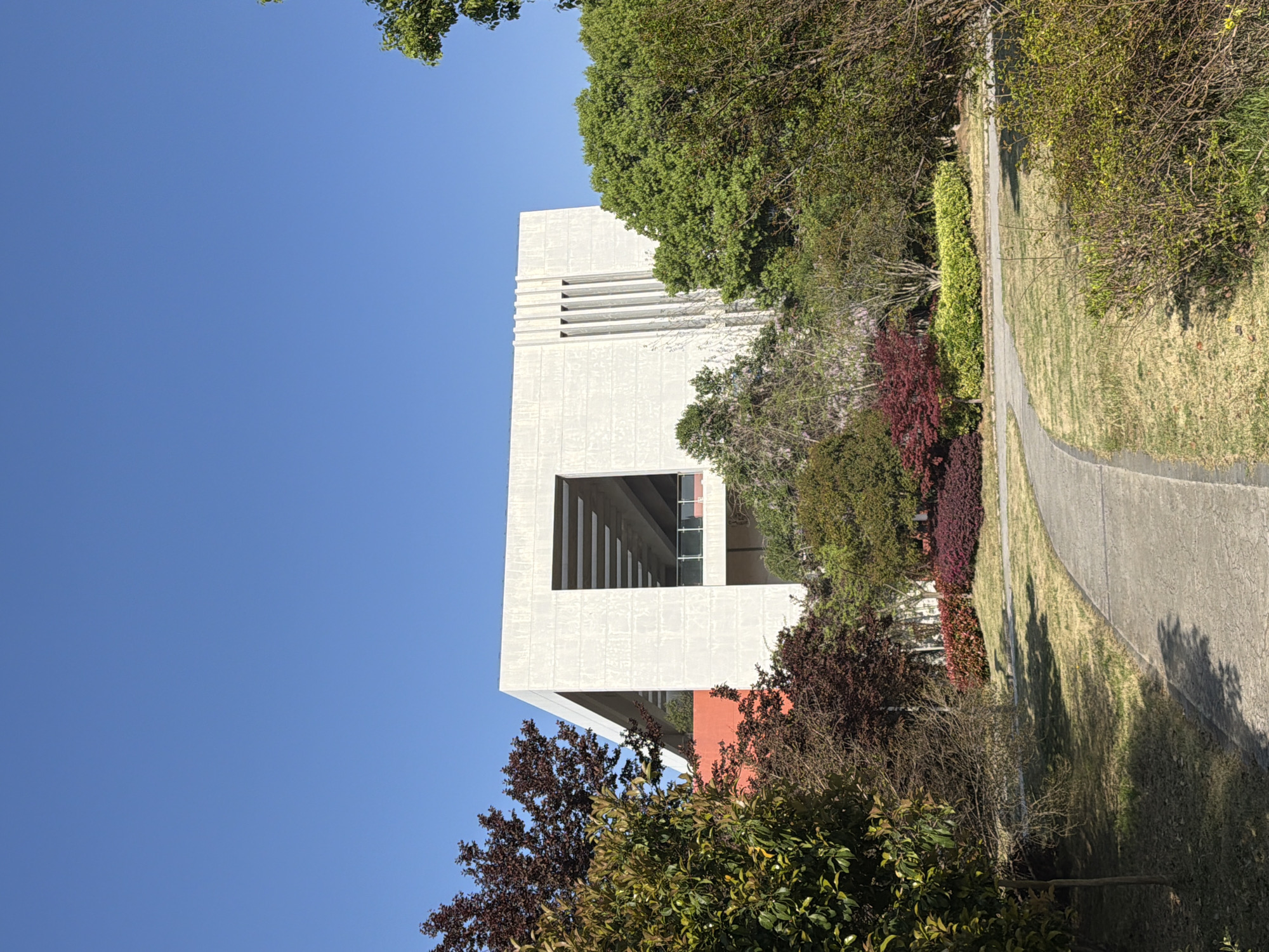 The image depicts a modernist, light-colored building with distinct horizontal recessed stripes near its top and a large rectangular opening. The building is framed by lush green trees and various bushes with red and green foliage. A paved pathway runs alongside the vegetation. A clear blue sky is visible above. The location is Jiangning District, China.