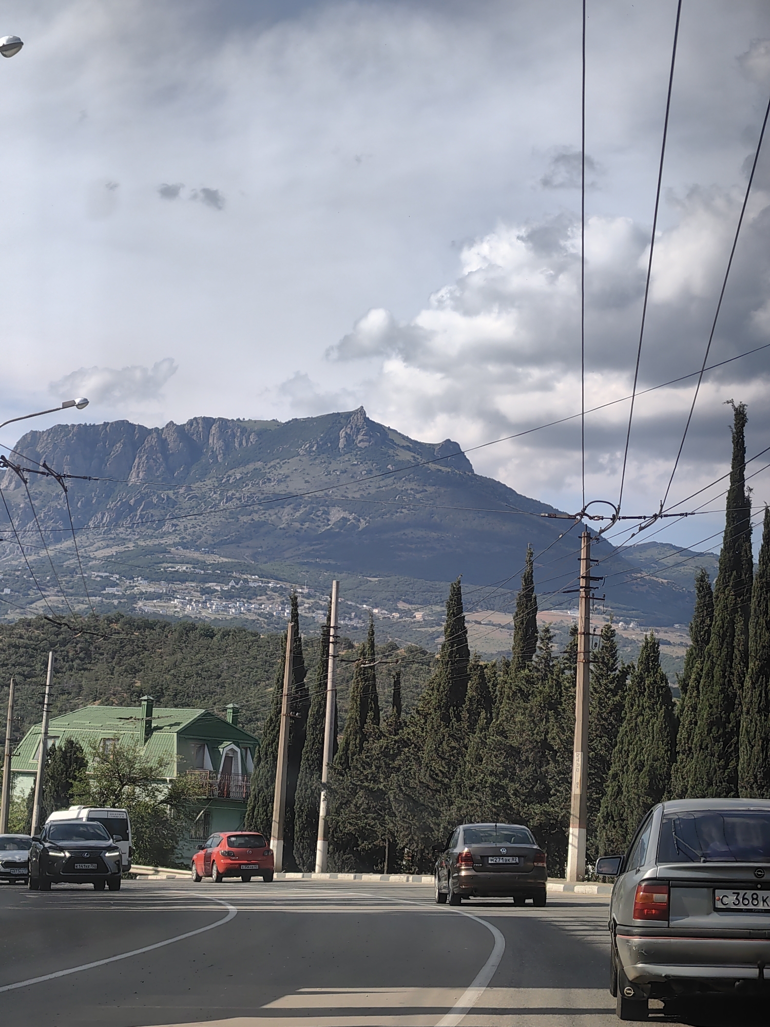 The image shows a view from a road in Alushta, Ukraine. In the foreground, there are several cars on the road, including a grey sedan and a red hatchback. To the left, a large green building with a distinctive roofline is visible, partially obscured by trees. In the background, a majestic mountain range dominates the landscape, with a small town or village nestled on its slopes. Tall cypress trees line the roadside, adding to the scenic beauty of the location. Overhead, power lines and poles crisscross the sky, indicating the presence of infrastructure. The sky is partly cloudy, with a mix of sunlight and shadows creating a dynamic atmosphere.