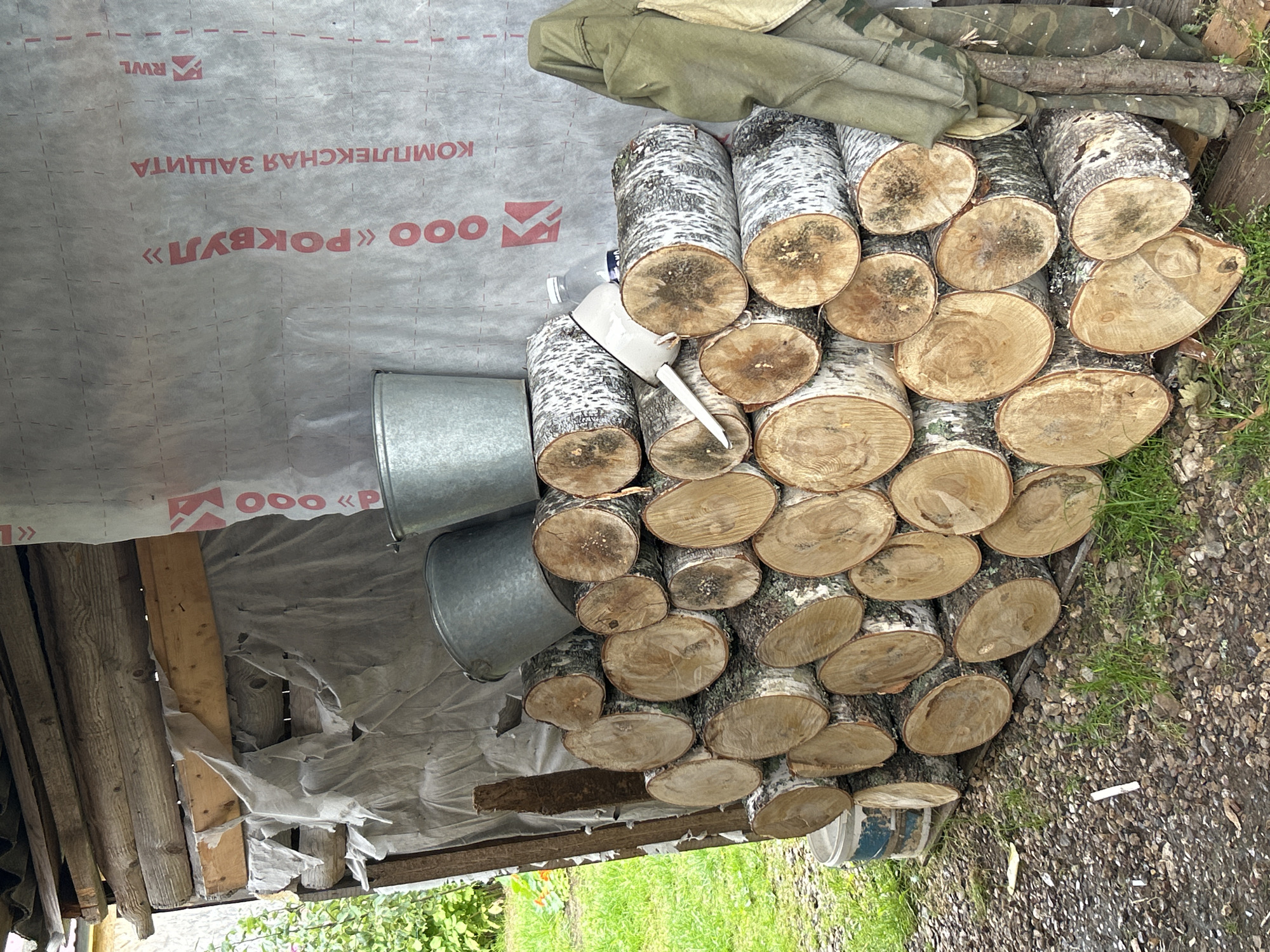 The image depicts an outdoor scene in the city of Kalitsino, Russia, characterized by a rustic aesthetic. In the foreground, a substantial pile of freshly cut birch logs is neatly stacked. The logs are of varying diameters and lengths, with their distinctive white bark and dark markings visible. Some logs show visible grain patterns and knots on their cut ends.
Behind the logs, a rough wooden structure is partially visible, constructed from rough-hewn logs. This structure appears to be covered with a translucent, greyish material, possibly a tarp or building wrap, bearing red lettering and logos, including "ООО «РОКВУЛ»" and "КОМПЛЕКСНАЯ ЗАЩИТА".
On top of the logs, two galvanized metal buckets are placed. To the right of the stacked logs, a piece of olive-green outerwear, possibly a jacket or coat, is hanging from a wooden post, along with a section of camouflage-patterned fabric. A small white plastic funnel is also visible amidst the logs.
The ground surrounding the logs is covered with a mixture of gravel and dirt, with sparse patches of green grass growing through. To the left of the logs, a blue and white bucket is partially visible. In the background, to the left, glimpses of green foliage and possibly a yellow flower can be seen, suggesting a natural outdoor setting. The overall impression is one of preparation for winter or a rural storage area.