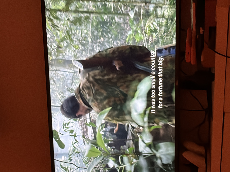 The image displays a television screen showing a cinematic scene, likely from a film or series, within a domestic setting.
**On the TV Screen:** A male figure, viewed from behind, is positioned slightly right of center. He wears a camouflage pattern jacket and has a dark-colored rifle with a wooden stock slung over his left shoulder. He appears to be moving through dense, lush green jungle or forest vegetation, with various plants and trees obscuring the background. Other indistinct figures and structures are faintly visible through the foliage in the distance. White subtitles are present at the bottom of the screen, reading: "It was too small a country / for a fortune that big."
**Off the TV Screen (Domestic Setting):** The television screen is situated on a light-colored wooden surface, likely a media console or cabinet. Below the screen, several items are visible: to the left, a light-colored, possibly fabric or soft plastic object with a curved shape; in the center, dark cables are coiled or resting; and to the right, a red flat object (possibly a book or tablet) rests on top of a dark electronic device, with another red object visible near it.
**Location:** The image provides no identifiable landmarks or contextual information to determine its specific location within the city of London, United Kingdom, or any other city. The scene displayed on the television screen depicts a jungle environment, and the domestic setting where the photograph was taken is generic.