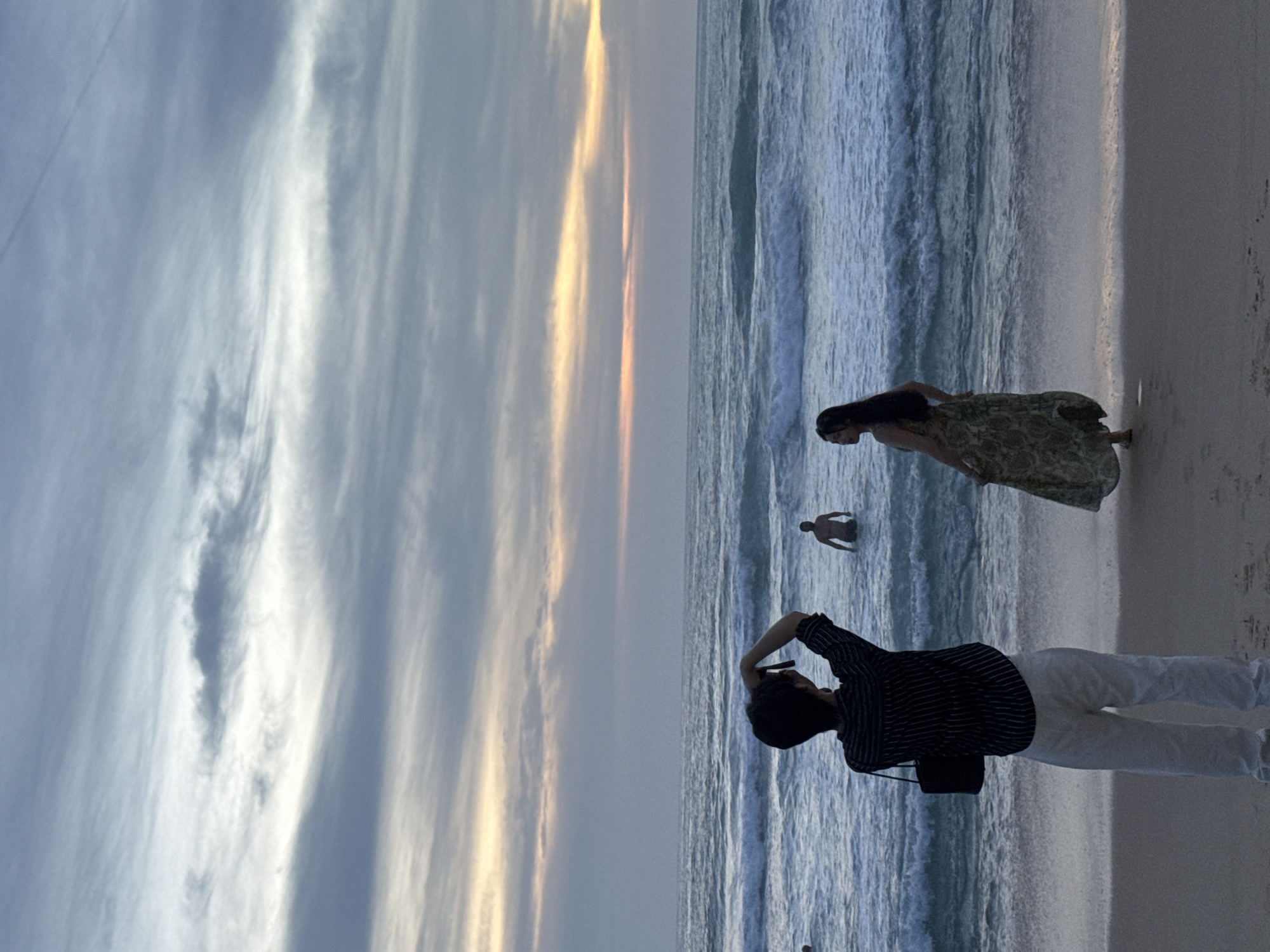 At Karon Beach in Karon, Thailand, a person wearing a striped shirt and white pants stands on the sand, facing the ocean and holding a camera up to take a photo. To the right of this person, a woman in a patterned dress walks along the shoreline. In the ocean, a man is partially submerged in the water, facing away from the shore. The sky above is filled with dramatic clouds, hinting at either sunrise or sunset.