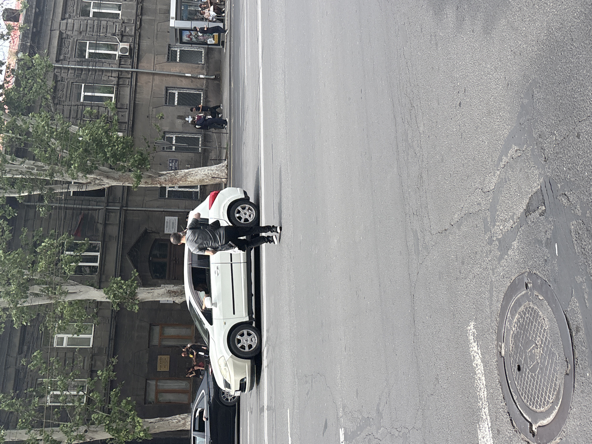 The image depicts a street scene in Yerevan, Armenia. The photograph shows a man standing beside a white car parked on the side of the road. Adjacent is a black car and an old dark building with multiple windows. Trees line the building. A manhole cover is visible on the right side of the image. Several people are standing on the side of the road.