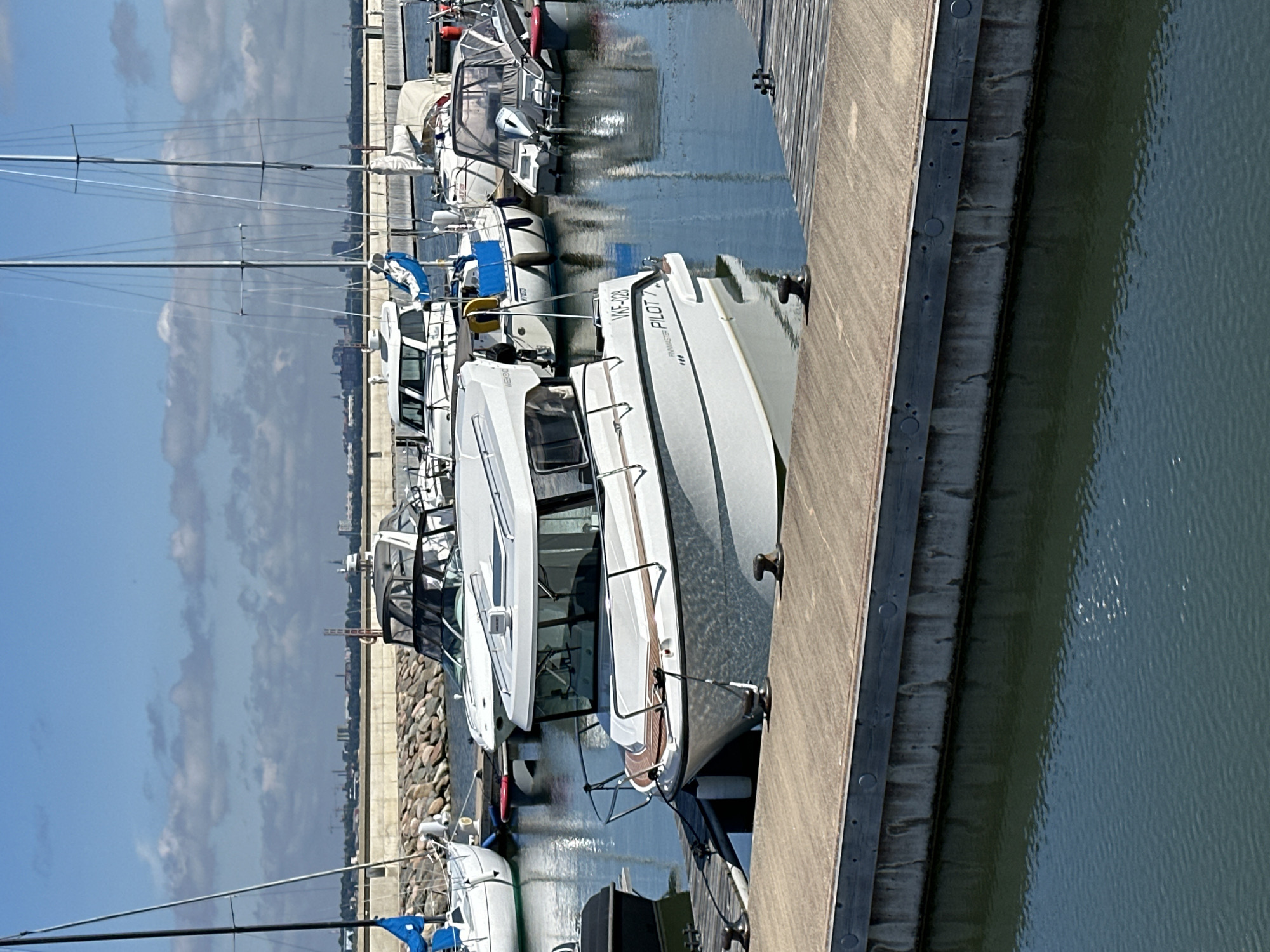 The image depicts a harbor in Tallinn, Estonia, on a sunny day with scattered clouds. In the foreground, a wooden dock extends into the calm, blue water, with mooring bollards visible. Several boats are docked side-by-side.
The most prominent boat in the foreground is a white motorboat with a dark trim around its windows and a light brown wooden deck area at the bow. It has a cabin with windows and a dark canopy. The boat is identified by the registration "VKF-028" and the text "PILOT" on its hull.
Behind this motorboat, other vessels are visible. To the left, a white sailboat with a blue sail is partially seen. Further back, there are more sailboats and motorboats, some with covers. In the background, a long concrete and stone seawall separates the harbor from a strip of land with buildings and cranes, suggesting an urban or industrial area. The cityscape of Tallinn is faintly visible in the distance. The overall scene conveys a peaceful marina environment.