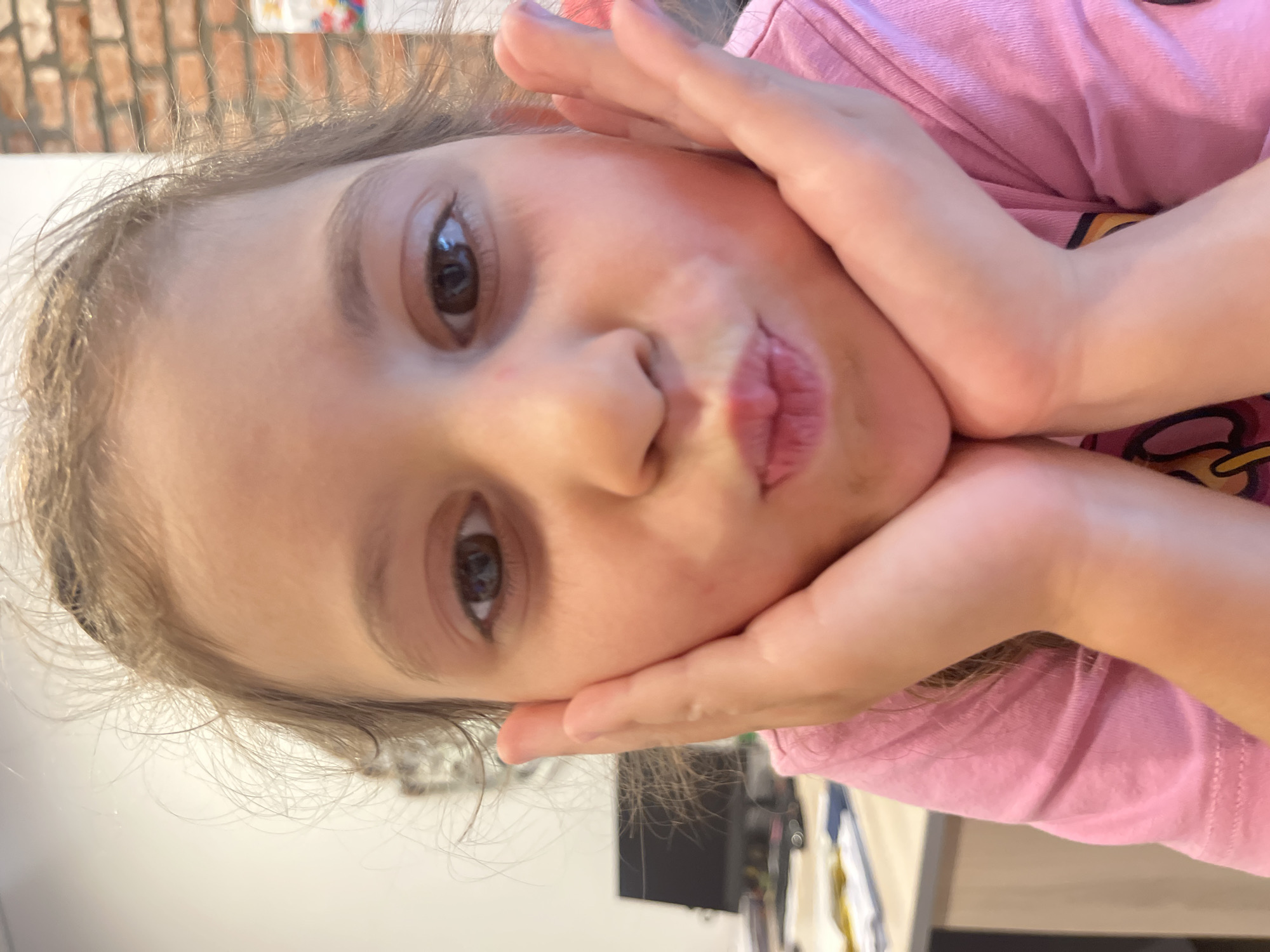 A close-up shot features a young girl with brown eyes and light brown hair resting her head on her hands. She is wearing a pink t-shirt. Her expression is somewhat playful, with her lips pursed. The background is slightly blurred, showing what appears to be a brick wall and some other indistinct objects. The lighting suggests it could be daytime. The image is oriented sideways.