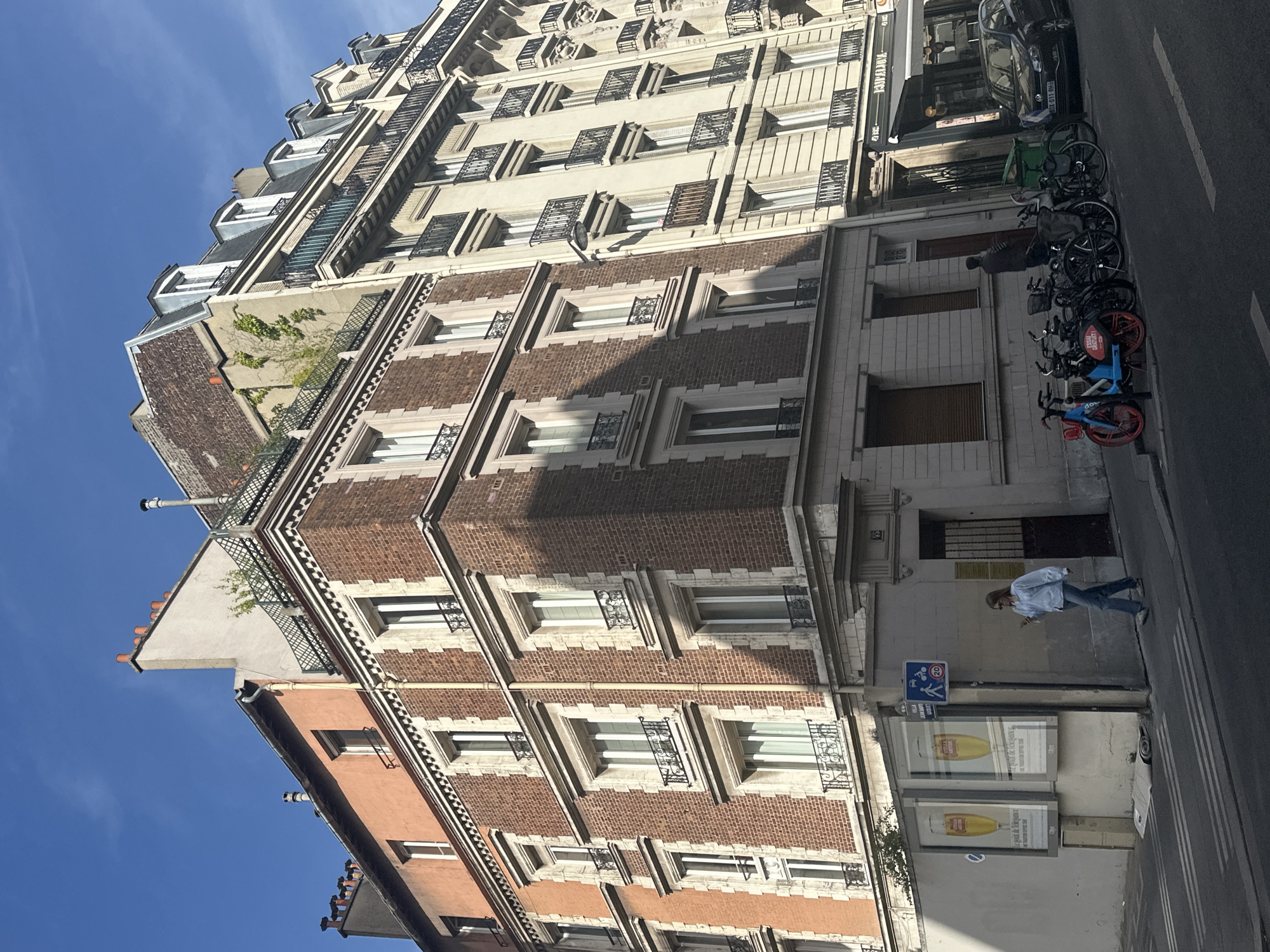 The image depicts a street scene in Paris, France, showcasing a row of multi-story buildings under a clear blue sky. The buildings are a mix of architectural styles, with the foreground featuring a brick building with ornate stone trim around its windows and several floors. Adjacent to it is a lighter-colored building with balconies and more windows. In the background, another building with a similar style and many windows is visible.
On the street level, a woman in a light blue shirt and jeans walks on the sidewalk, looking at her phone. In front of the buildings, there are several parked bicycles, including a distinctive blue and red bike-share bicycle. There are also advertisements displayed on a wall and a dark-colored car parked on the street. The scene appears to be a typical urban environment in Paris, with the characteristic Haussmannian architecture and street amenities.