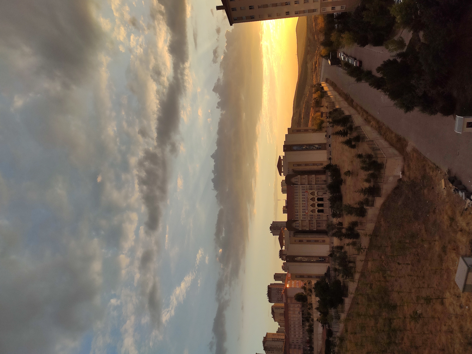 The image captures a sunset view over a cityscape, likely in Etimesgut, Turkey, characterized by apartment buildings and a large, institutional-looking structure. The sky is filled with a mix of dark and light clouds, with rays of sunlight piercing through, casting a warm glow on the horizon.
In the foreground, a somewhat barren hillside slopes down towards a road. Young trees are sparsely planted on the hillside. A concrete wall runs along the edge of the property. Several apartment buildings are visible, with the largest structure in the center appearing to be a school or university building, featuring classical architectural elements like arched windows and columns. To the right, a tall apartment building stands prominently.
A paved road runs diagonally through the mid-ground, with several parked cars visible along its side. The surrounding area is dotted with trees, some of which appear to be evergreens. In the background, rolling hills are illuminated by the setting sun. The overall impression is of a developing urban or suburban area at dusk.