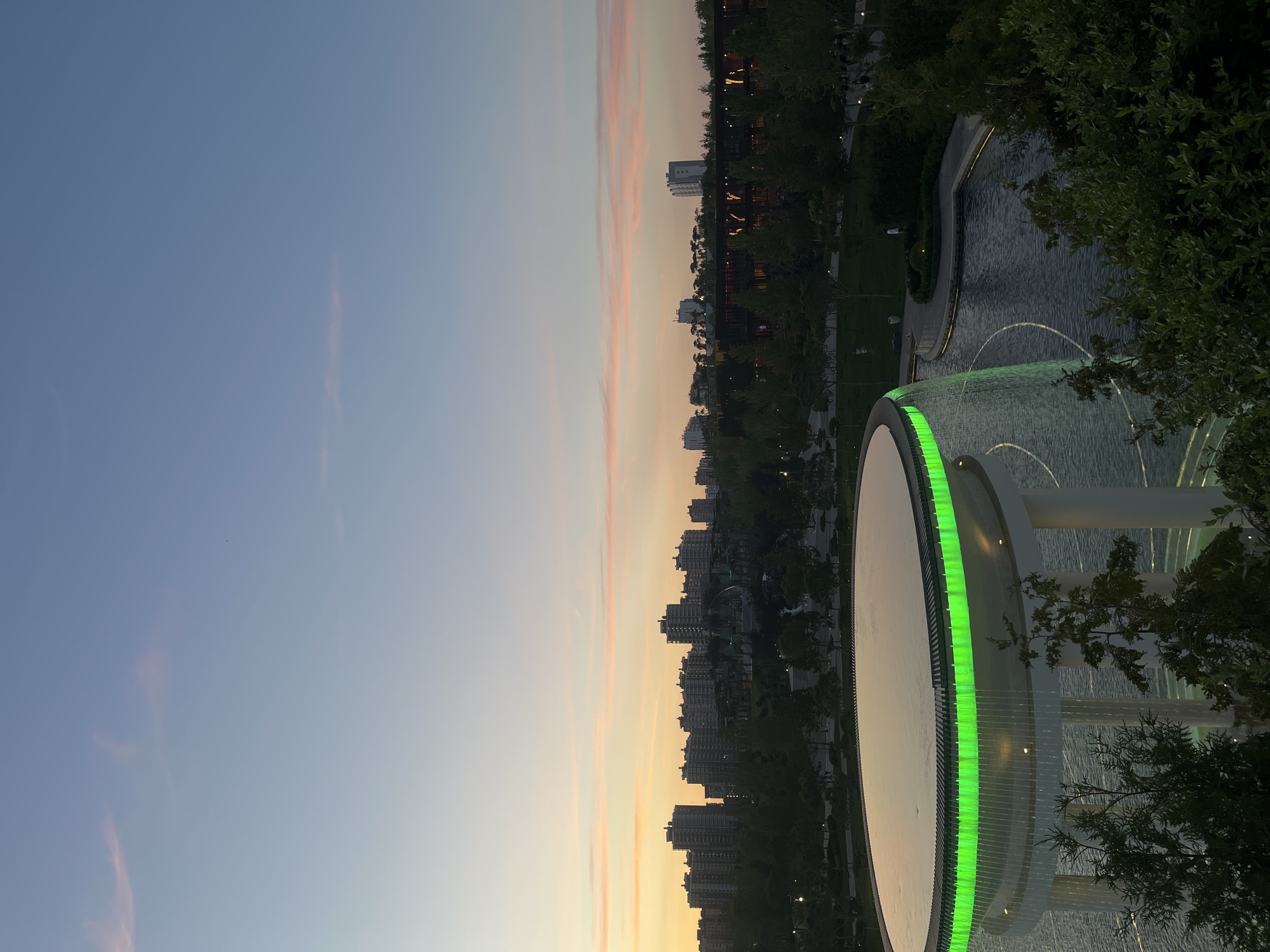 This is an outdoor, eye-level shot taken at dusk in Krasnodar, Russia, likely in the "Krasnodar" Park, formerly known as the "First Hundred Years of Krasnodar" park. In the foreground, a round, elevated platform with a rim of bright green lights is visible. Water cascades over the rim, forming a curtain of falling water. Beyond the platform, a body of water, possibly a pond or lake, is present, with circular light patterns visible beneath the surface. In the midground, a park landscape with trees and pathways unfolds. In the background, a cityscape of multi-story residential buildings and some commercial structures stretches across the horizon. The sky above is a gradient of soft blue and orange, indicating the setting sun, with faint pink clouds. Several people can be seen walking in the park and on the pathways, appearing as small, indistinct figures. The overall scene conveys a sense of urban recreation and twilight ambiance.