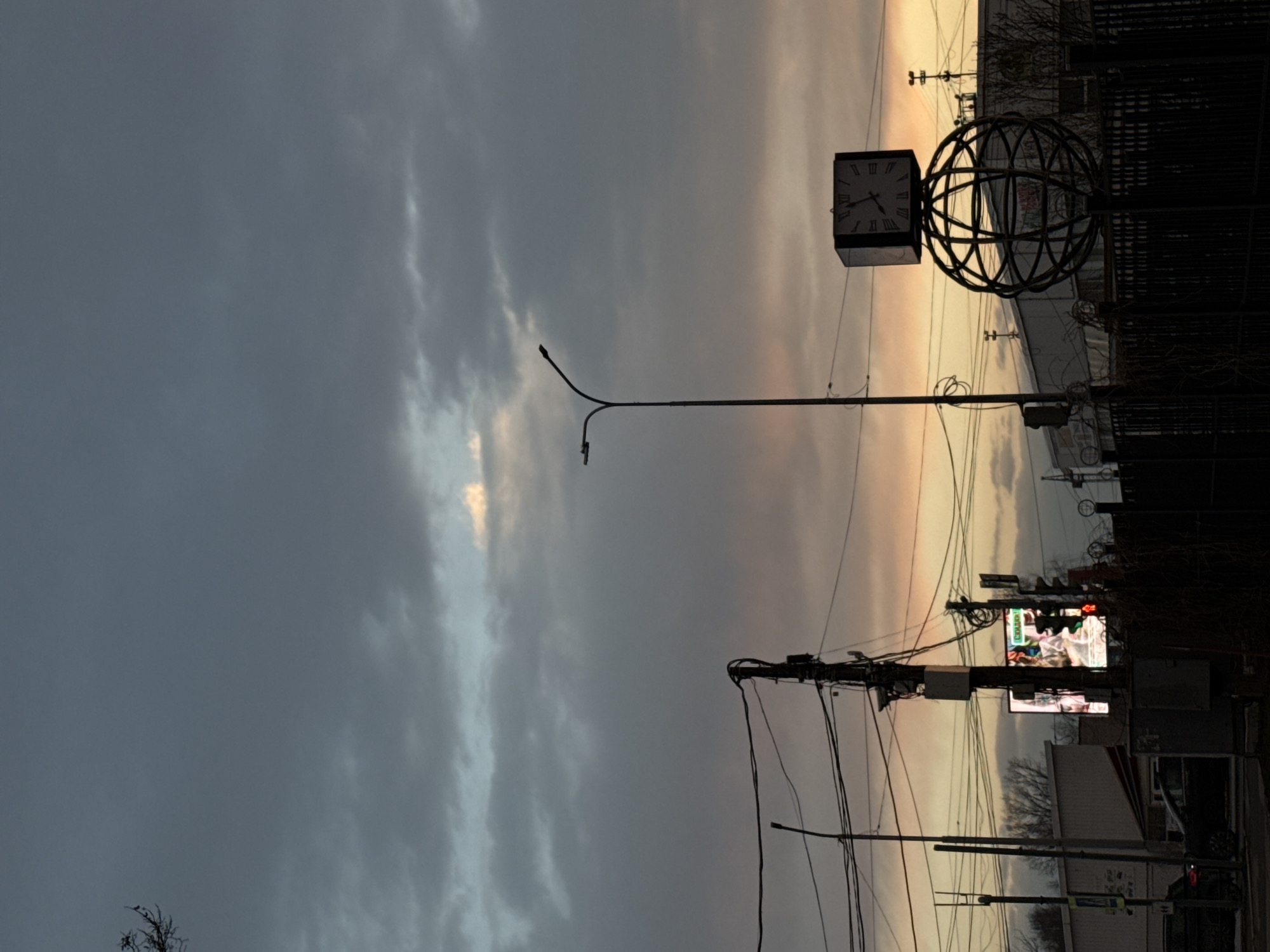 The image depicts an evening cityscape in Chaikovsky, Russia, under a sky with dramatic cloud formations ranging from dark grey to pale orange at the horizon. Dominating the upper right is a large, metal street clock with Roman numerals, supported by a metal globe-shaped structure, set against a building or fence. A street lamp is visible extending towards the left. Below and to the right, power lines intersect with traffic signals, street lighting, and advertising displays atop buildings along a street with limited visibility. Minimal details suggest a vehicle is present on the street.