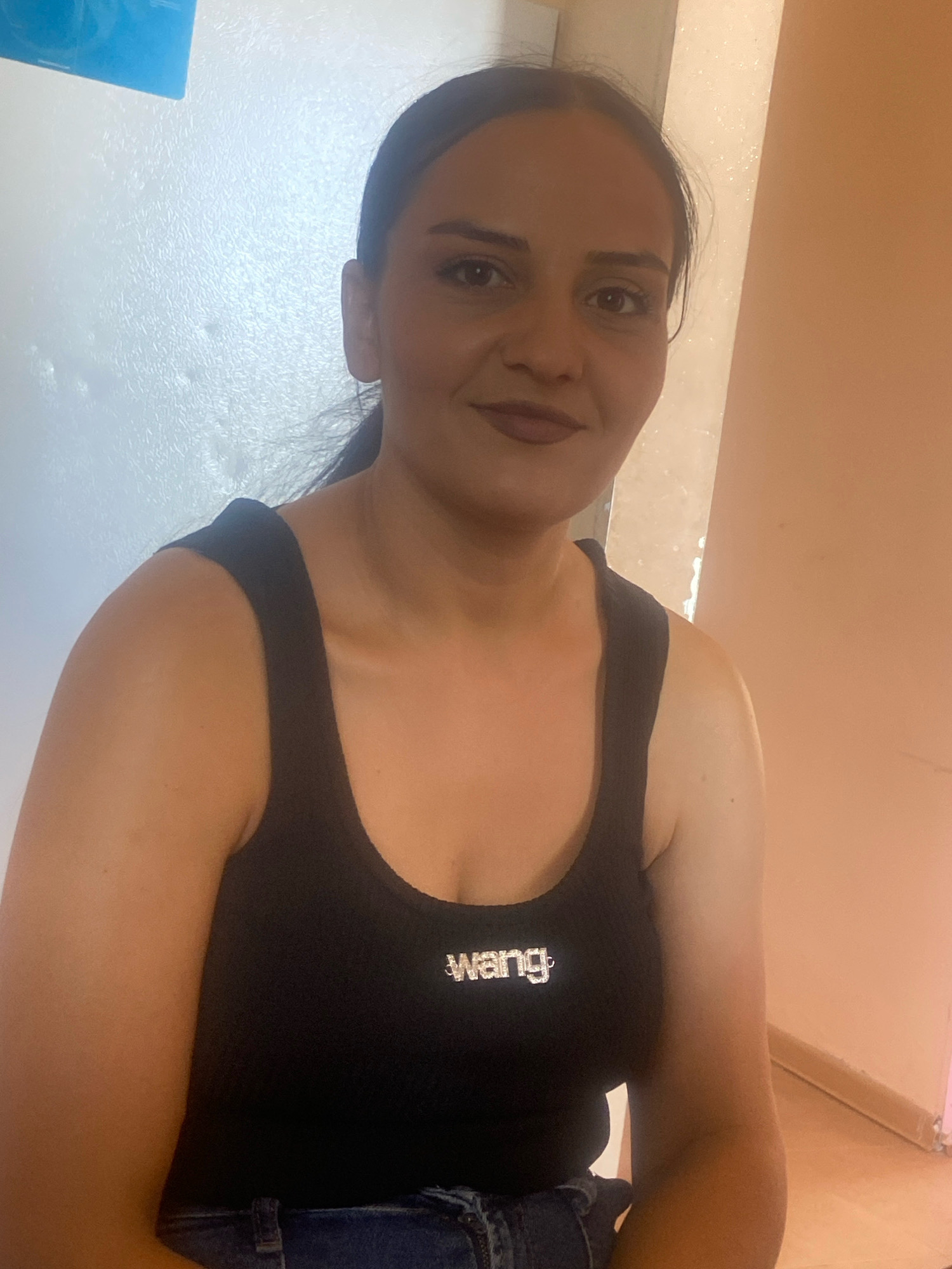 A woman with dark hair pulled back in a ponytail wears a black tank top with "WANG" in rhinestones across the chest. She has on dark denim jeans. The woman is seated indoors with a textured white wall behind her left side and a peach-colored wall behind her right. A faint blue object is visible in the upper left corner. The lighting is warm and casts shadows. The scene appears to be a casual indoor setting, possibly a home or a studio, in Nor Kharberd, Armenia.
