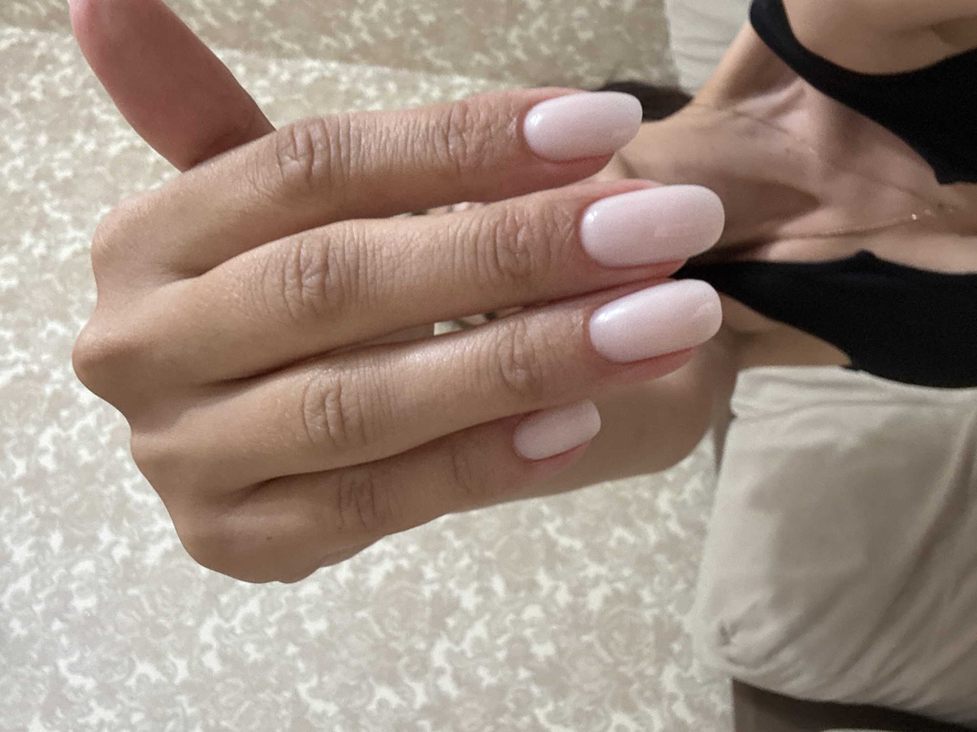 A close-up shot depicts a person's left hand with manicured fingernails painted a light pink color. The hand is held in a partially flexed position against a background of a light beige and white patterned fabric. Visible behind the hand is the person's upper chest and neck, dressed in a black top, with a gold necklace. There is no evidence in the image to suggest the location is in Sochi, Russia.