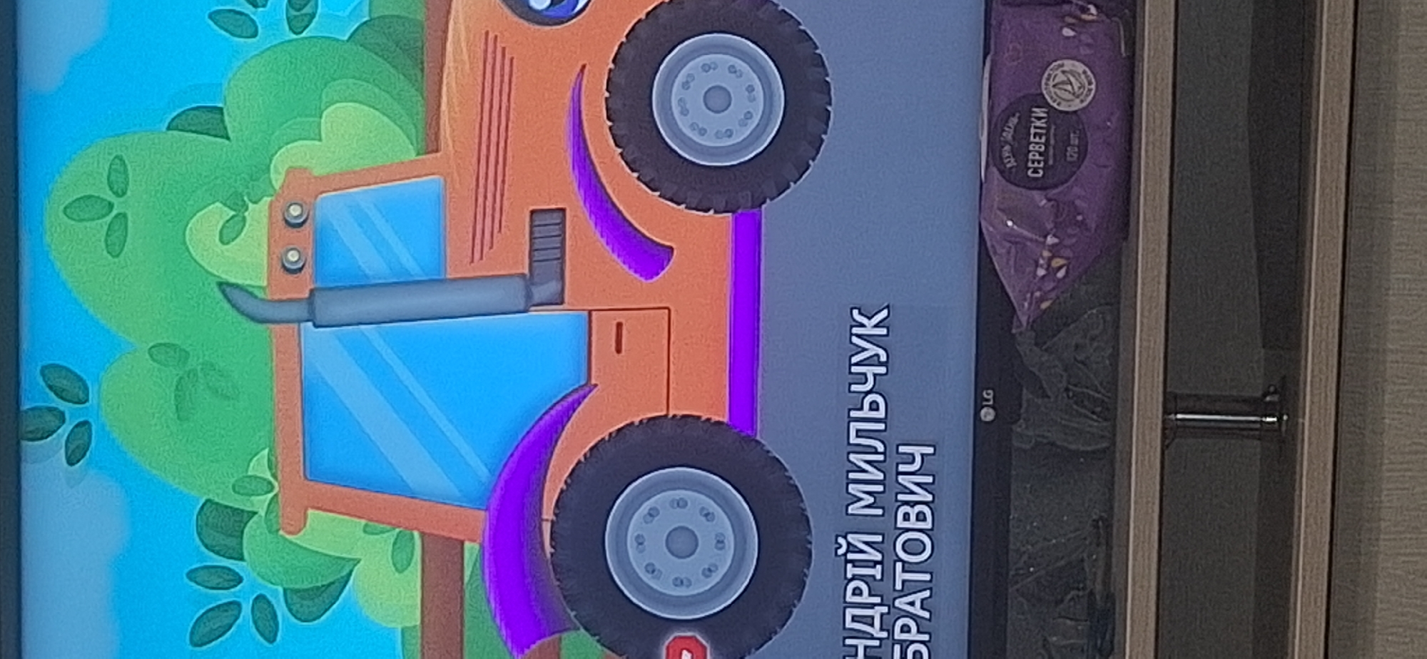 The image shows a close-up of a television screen displaying a cartoon image of an orange tractor with purple accents against a green, hilly background and a blue sky. Below the cartoon, text in white lettering reads "АНДРІЙ МИЛЬЧУК" and "БРАТОВИЧ". To the right of the text, the LG logo is visible. Below the television, a shelf holds a package of purple wet wipes with text in Russian that appears to say "СЕРБЕТКИ" (servetki, meaning wet wipes). The overall scene appears to be indoors, with the television and shelf set up in a room. There is no discernible indication of the location being in Bila Tserkva, Ukraine.