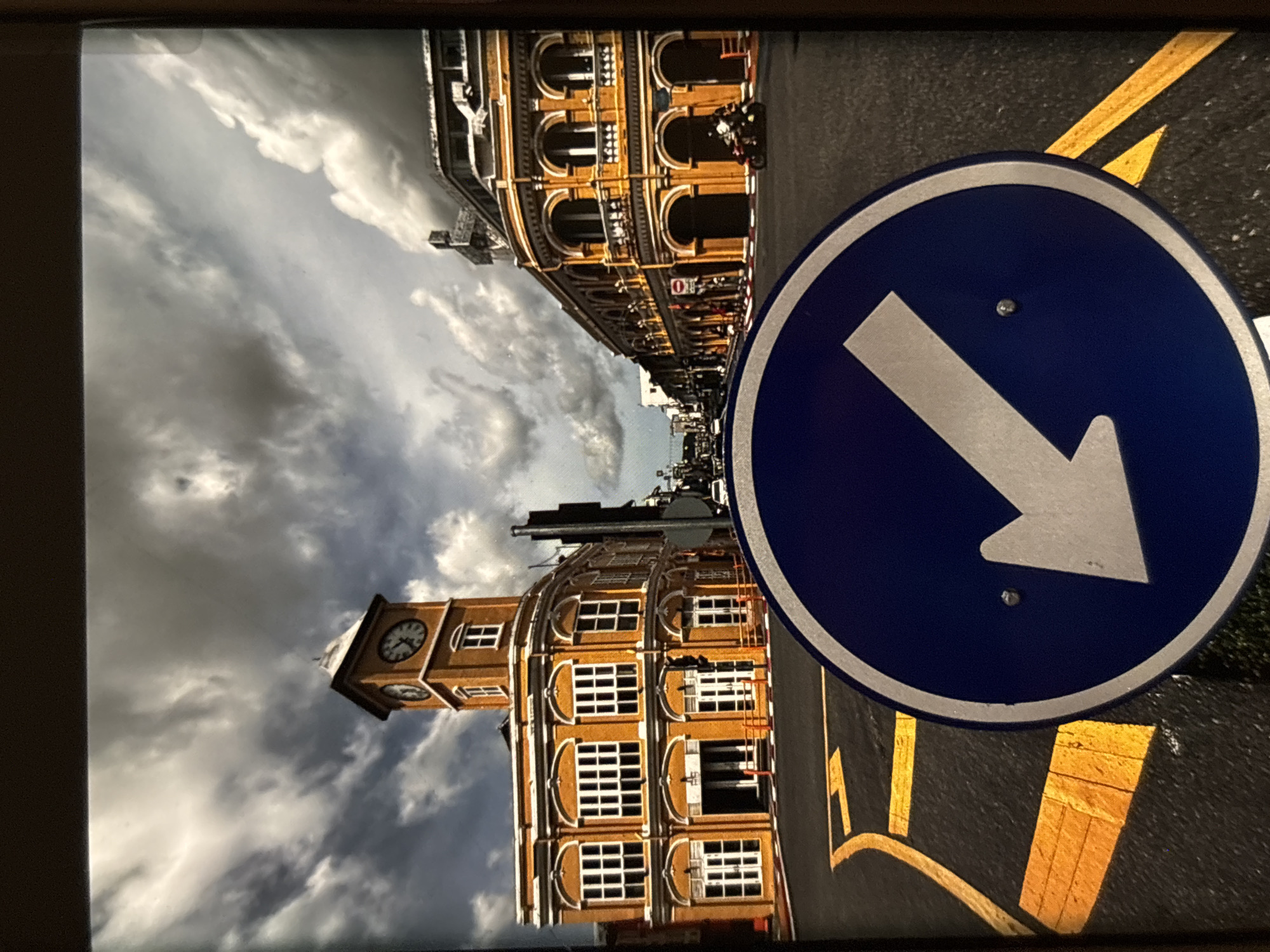 The image shows a street scene in Chalong, Thailand, with two prominent colonial-style buildings painted in bright orange. The building on the left features a clock tower. The sky above is dramatic with dark, heavy clouds contrasting with patches of lighter sky. In the foreground, a blue circular traffic sign with a white arrow pointing downwards and to the left is visible. The street is paved and has yellow lane markings. In the background, there are people, a motorcycle, and other vehicles, suggesting an active urban environment. The overall impression is of a bustling street scene under a moody sky.