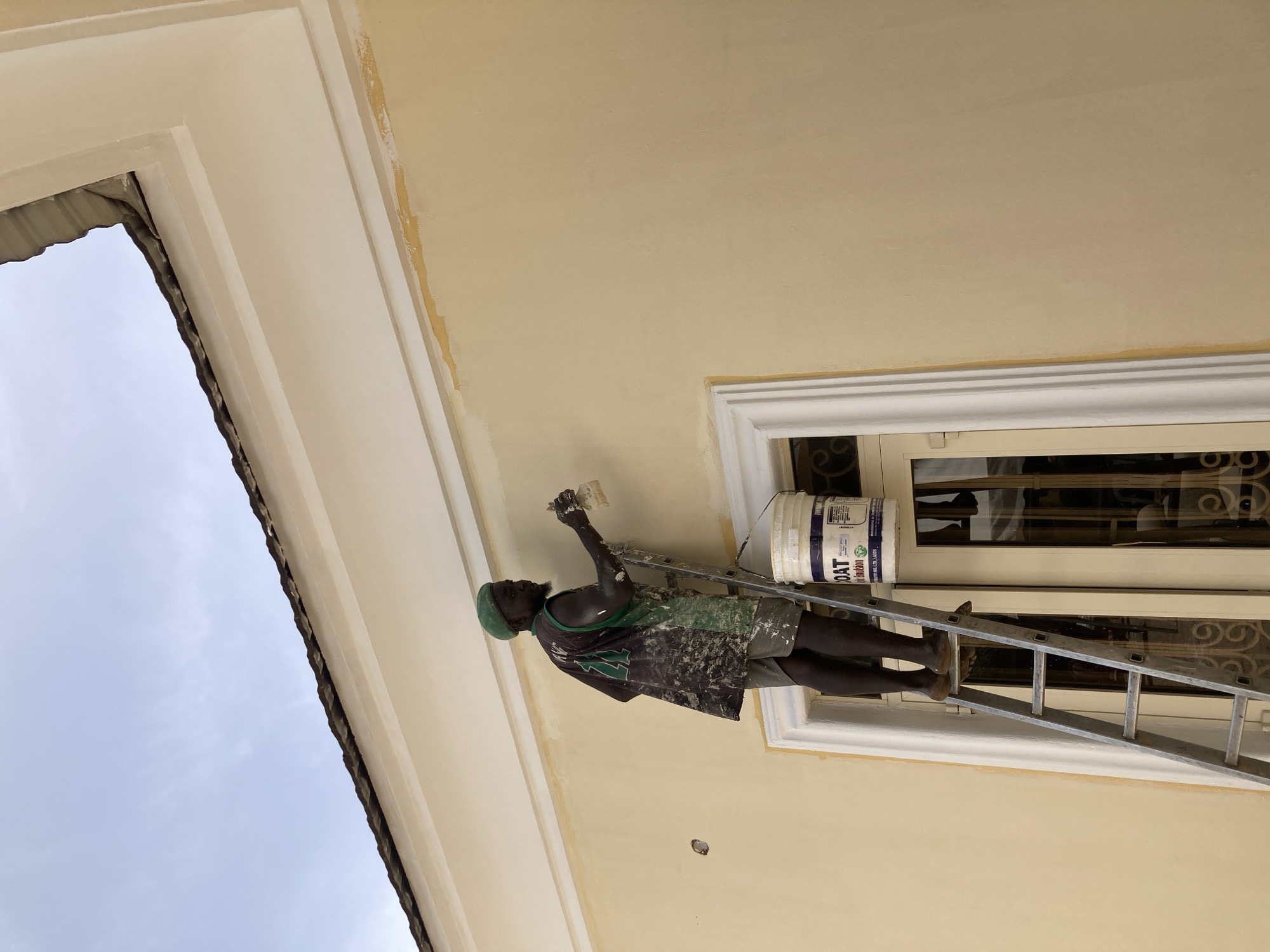 An exterior shot shows a man painting a building wall in Jos, Nigeria, under a bright, overcast sky. The man, with dark skin, wears a green beanie, a paint-splattered t-shirt (dark green, black, white pattern), and dark shorts. He stands barefoot on a silver extension ladder, positioned against the wall next to a window.
He holds a paintbrush in his right hand, actively applying a lighter, creamy beige paint to the upper part of the wall, which is currently a similar but slightly darker or less uniform cream color. Above the window, to his right, rests a white plastic paint bucket. Visible text on the bucket includes "BOAT EMULSION" and "MANUFACTURED BY PREMIER PAINTS INDUSTRIES LTD, LAGOS," along with a green leaf logo and other small print.
The building features a multi-layered, white decorative trim around the window and along the roofline. The window itself has ornate, dark-colored ironwork. To the left of the painter, the edge of the roof is visible against the light grey sky. There are some splatters of paint on the ladder and the man's clothing, indicating ongoing work. The scene captures a moment of active construction or renovation work during daylight hours.