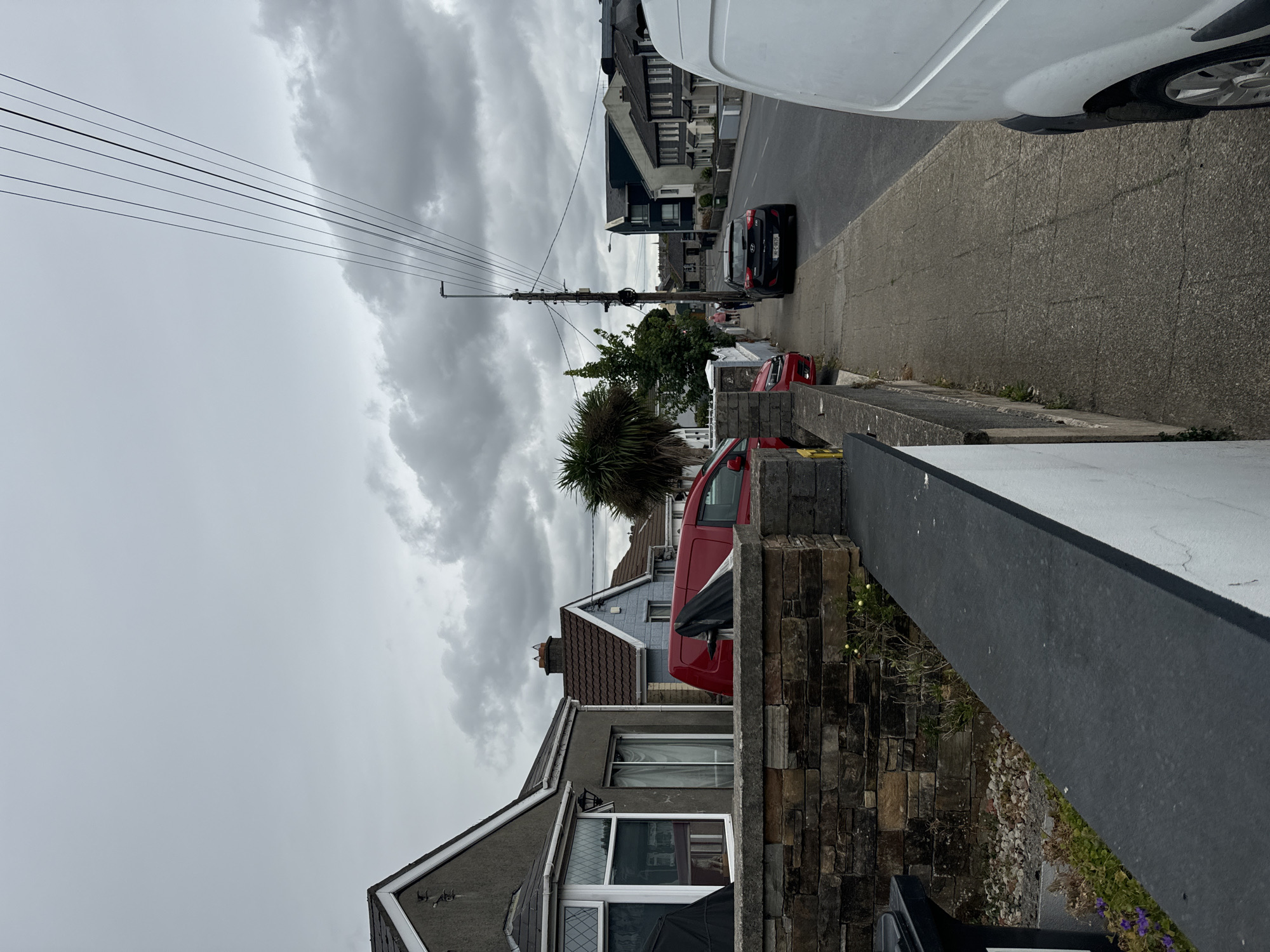 The image depicts a suburban street scene in the city of Waterford, Ireland, under a cloudy sky. On the left, a stone wall borders a property with a gray house featuring a bay window and a dark roof. Adjacent to it is a narrower gray house with a sliding glass door. A red van is parked in front of the houses. Further down the street, a blue house with a red-tiled roof is visible.
The sidewalk runs alongside the street, with a power pole and overhead wires dominating the center of the frame. A black car is parked on the street, and a white van is parked on the right side of the image, partially obscuring the view of the street and sidewalk further down. Two people are walking on the sidewalk in the distance. The street appears to be a residential area with houses lining both sides.
