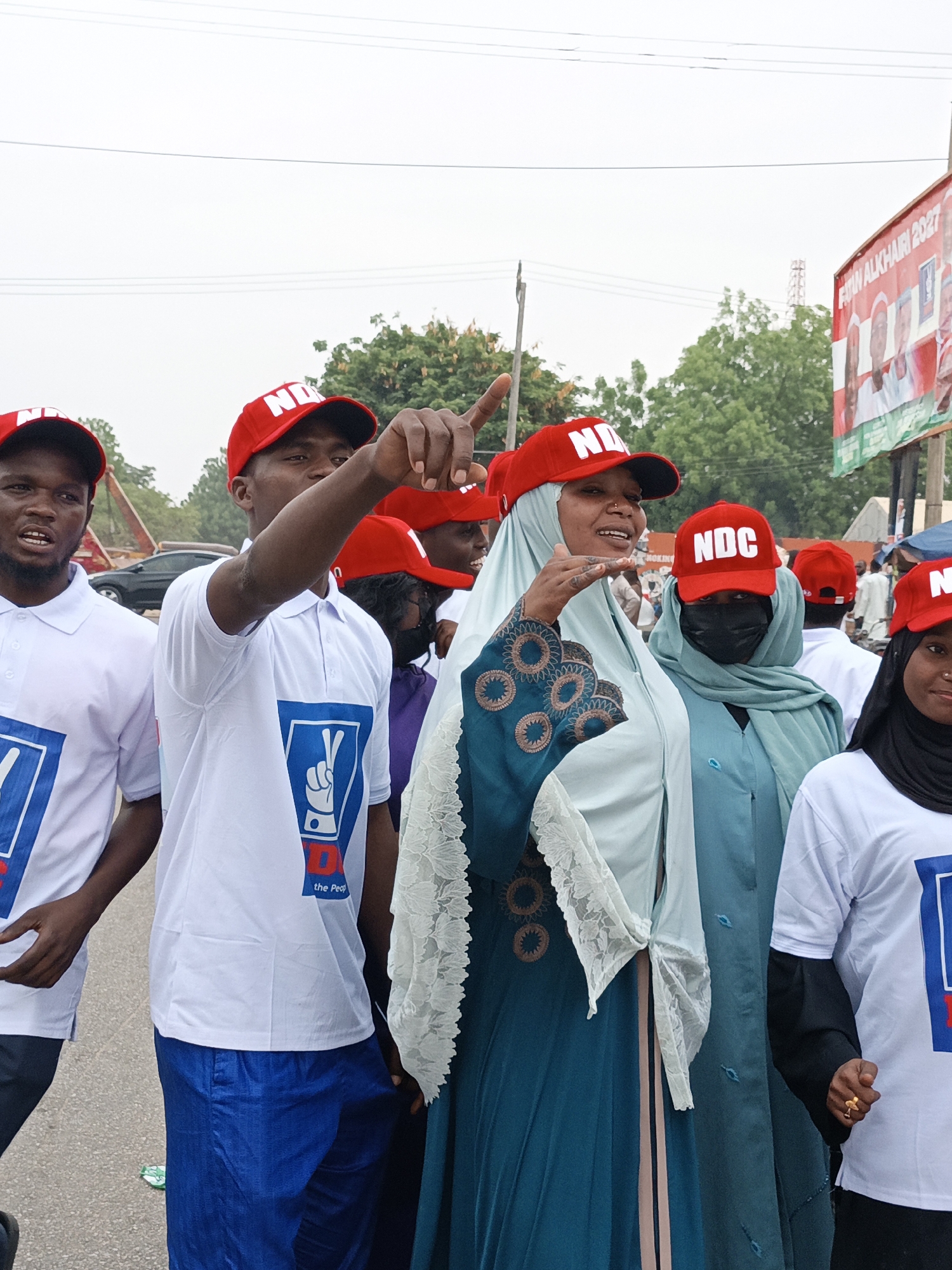 A medium shot captures a group of people in what appears to be a rally or campaign event, likely in the city of Tudun Wada, Nigeria. The attendees are wearing red caps marked with "NDC," and white t-shirts with a graphic of a hand making a "V" sign over the words "NDC the People." One man on the left side of the frame is pointing to the upper right; another man beside him is similarly pointing. A woman in a turquoise gown and pale blue hijab is gesturing with her hand. In the background, a billboard features images of political candidates, with the words "AMIN ALKHAIRI 2027." The setting seems to be an outdoor location along a street or road. The presence of trees and vehicles in the background suggests an urban environment.