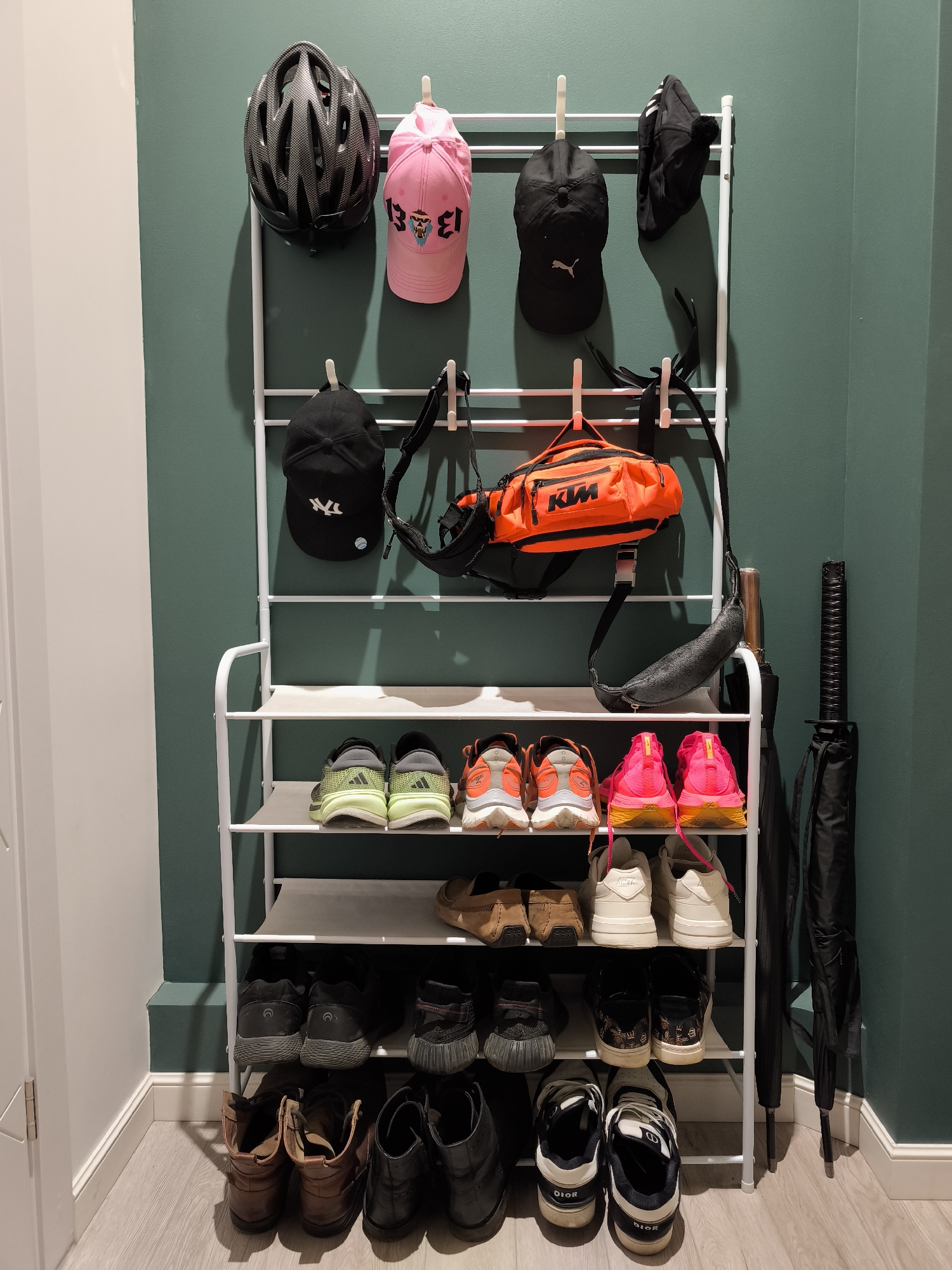 This image depicts a white, multi-tiered metal storage unit functioning as both a coat/hat rack and a shoe organizer, situated in an interior room in Moscow, Russia. The unit is placed against a smooth, matte-finish dark teal or green wall. A white door frame and an adjacent white wall section are visible on the left, with white baseboards lining the bottom of the walls. The floor consists of light-colored wooden laminate planks. The upper section of the storage unit features horizontal bars with white plastic hooks. From left to right on the top row, a black cycling helmet with a textured shell hangs from a hook, followed by a pink baseball cap with a stylized graphic (possibly "BDB" or "BOB") and a face emblem. Further right is a black baseball cap with a white Puma logo, and a black knit beanie with a white pom-pom and three white stripes. On the row below, from left to right, a black baseball cap with a white New York Yankees "NY" logo hangs. Next is a bright orange fanny pack or sling bag with "KTM" printed in black on its front, suspended by its black strap. To its right, a black patterned sling bag also hangs by its strap, which extends downward. The lower section of the unit comprises three shelves with grey fabric bases, designed for shoes. On the top shelf, a pair of grey and lime green Adidas athletic shoes are on the left, and a pair of orange and white athletic shoes are on the right. The middle shelf holds a pair of light brown suede loafers on the left, a pair of white athletic shoes with light brown soles in the center, and another pair of white athletic shoes on the right. The bottom shelf contains a pair of black athletic shoes with textured soles on the left, followed by a pair of black sneakers with white soles and intricate side patterns in the center, and a pair of black and white Dior branded athletic shoes on the right. On the floor directly beneath the unit, from left to right, are two pairs of brown leather boots (one lighter, one darker) and two pairs of black leather boots. To the far right, standing upright against the wall and the organizer, are two black umbrellas; the left umbrella features a katana-style handle. The scene is illuminated by an unseen light